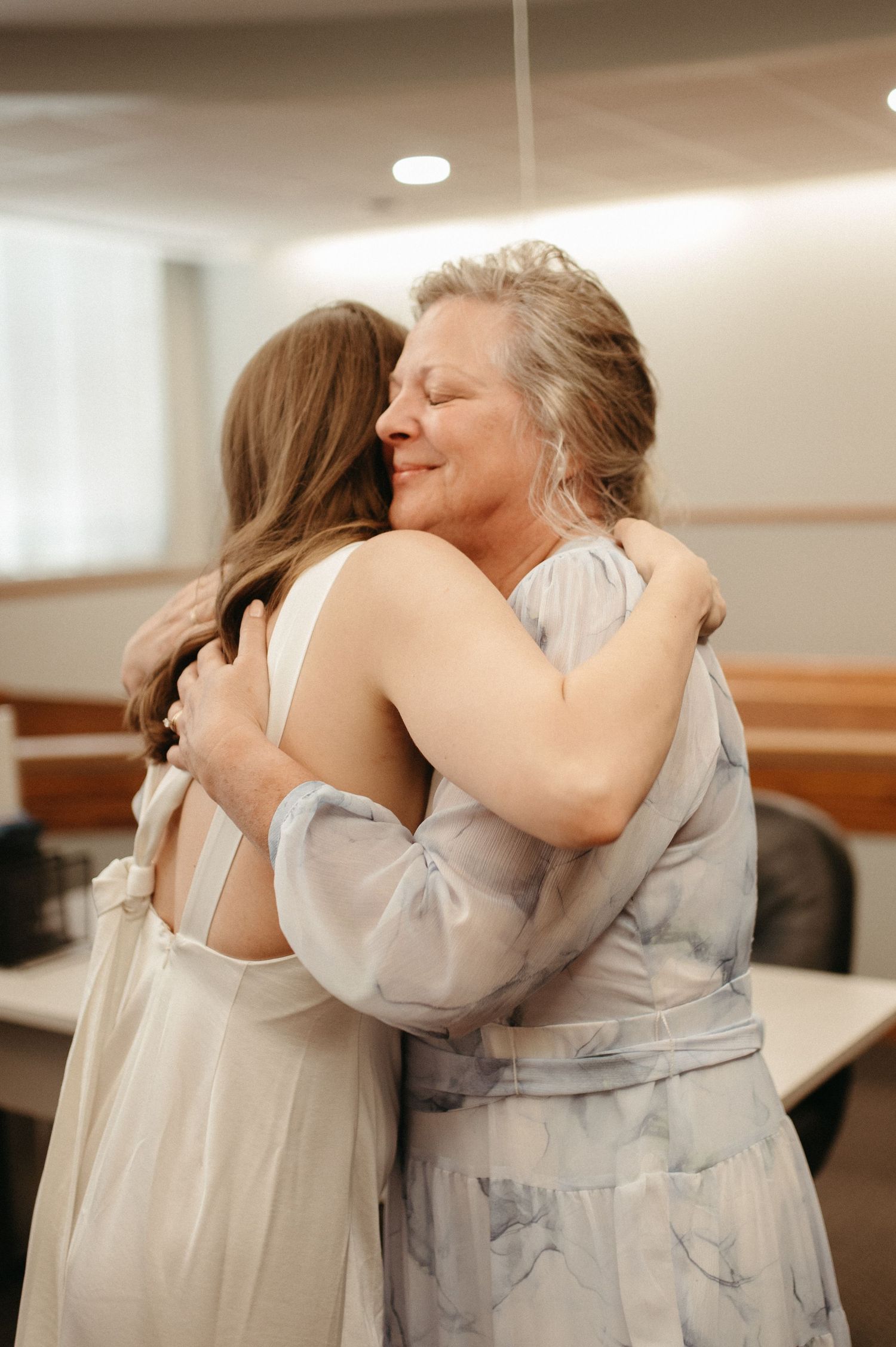 A warm hug between two women, one in a white dress and another in a patterned gray dress.