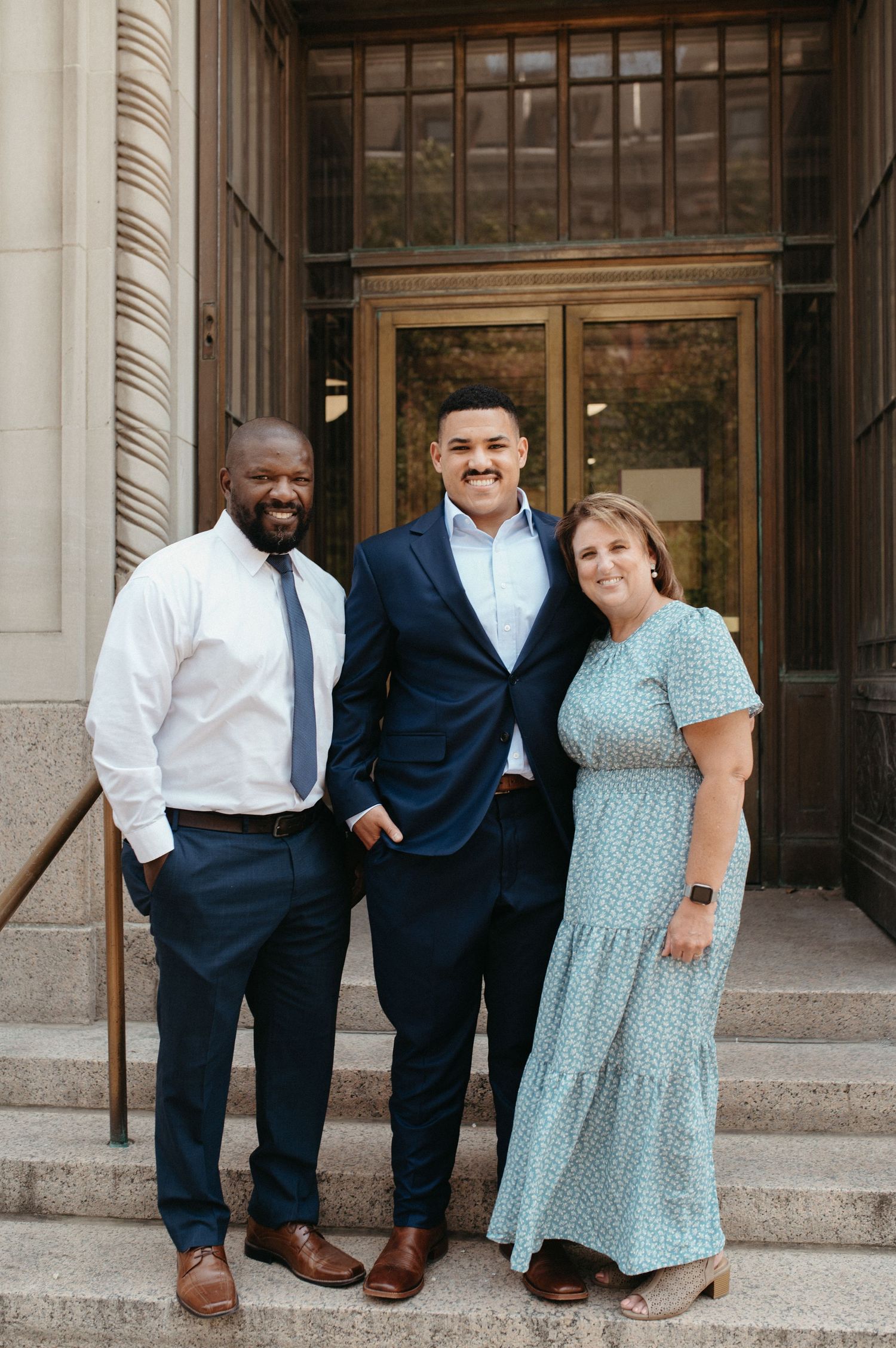 Three people in formal attire pose on stone steps in front of wooden doors.