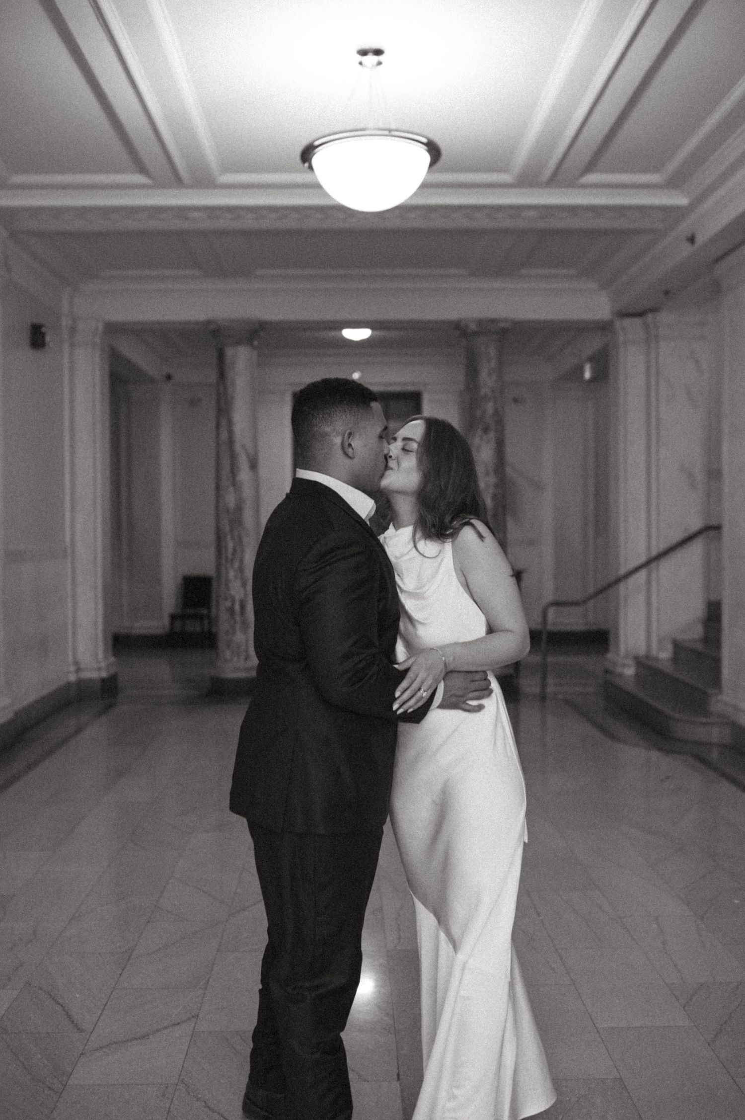 An intimate black and white photograph of a couple embracing in an elegant corridor.