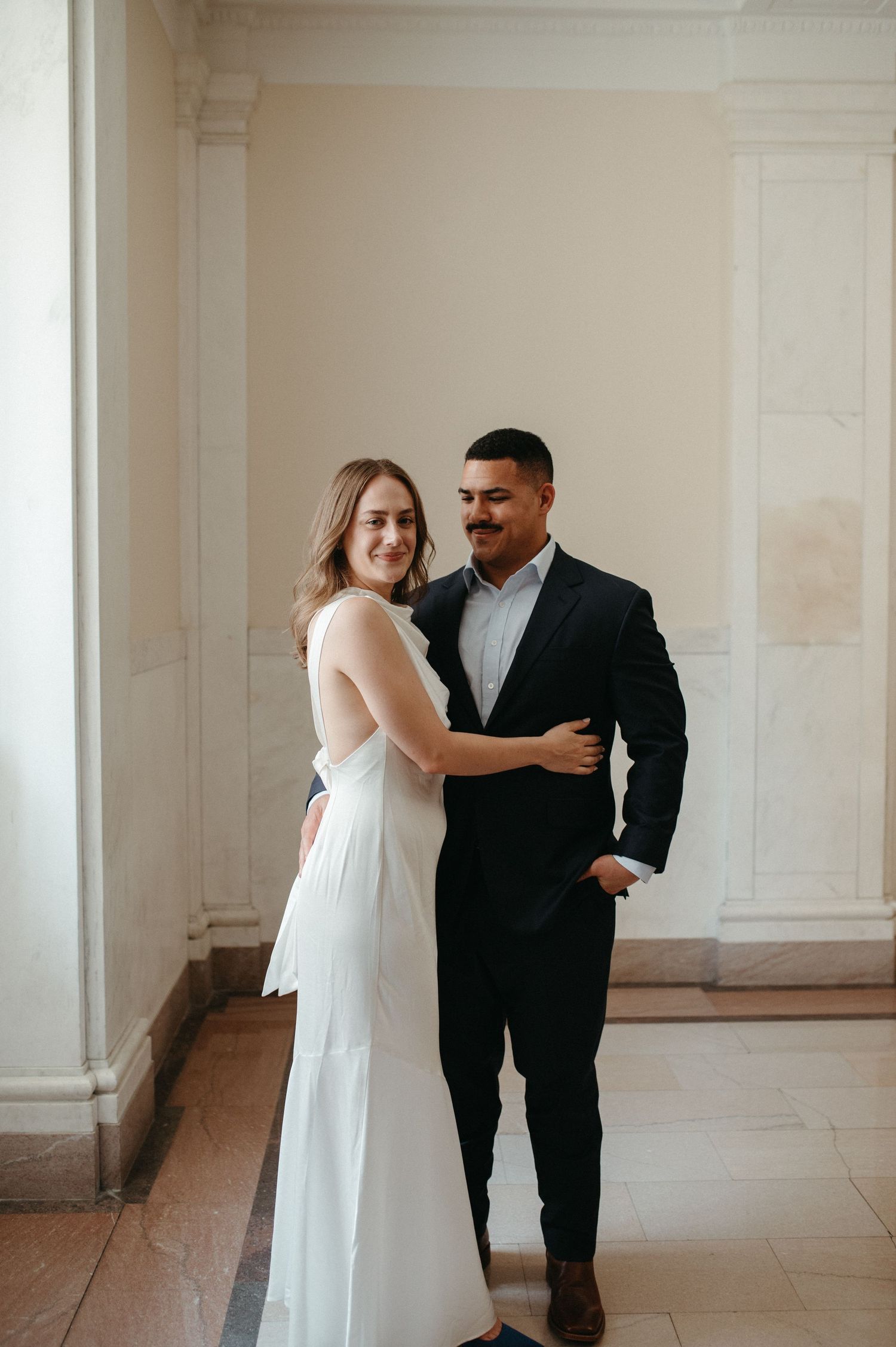 An elegant couple poses together in a bright hallway, the bride wearing a flowing white gown.