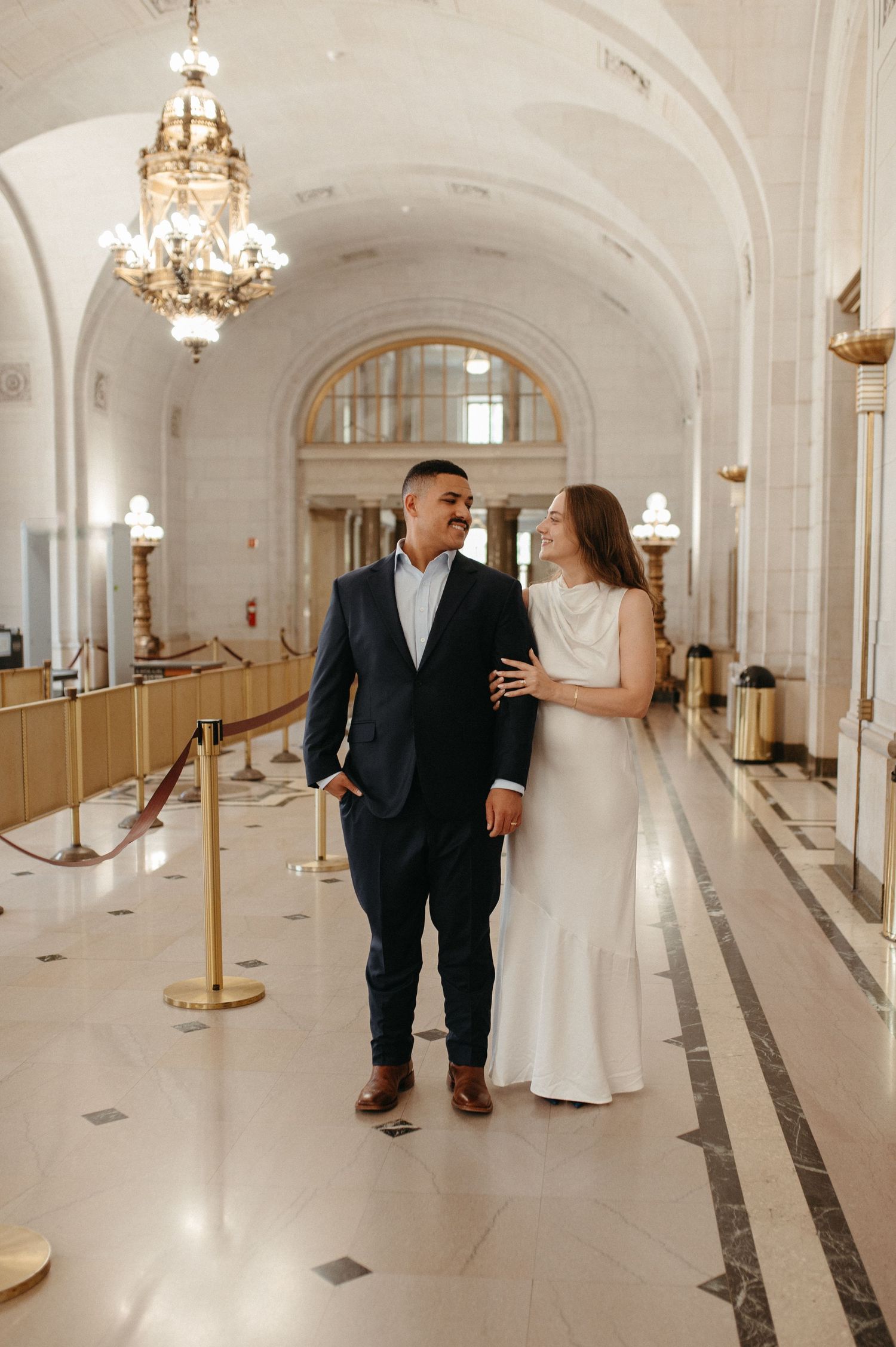 Couple strolls through grand hallway with ornate chandeliers and cream architectural details.
