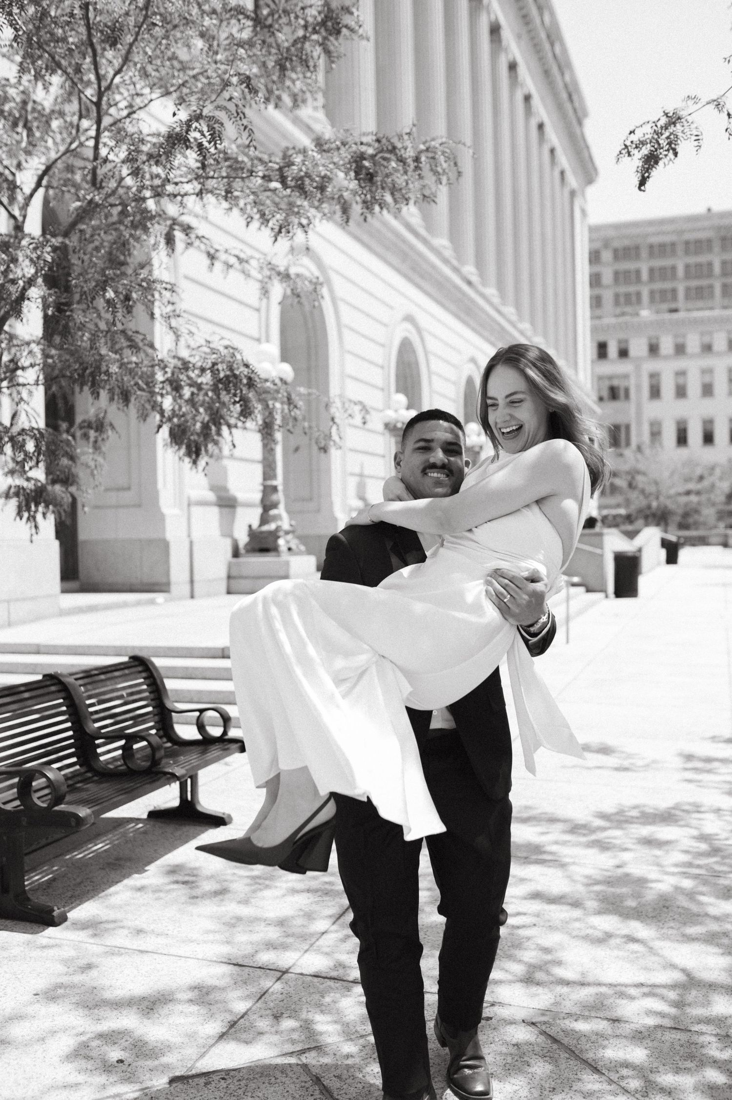 Groom lifts bride in white dress in front of classical building with park bench nearby.