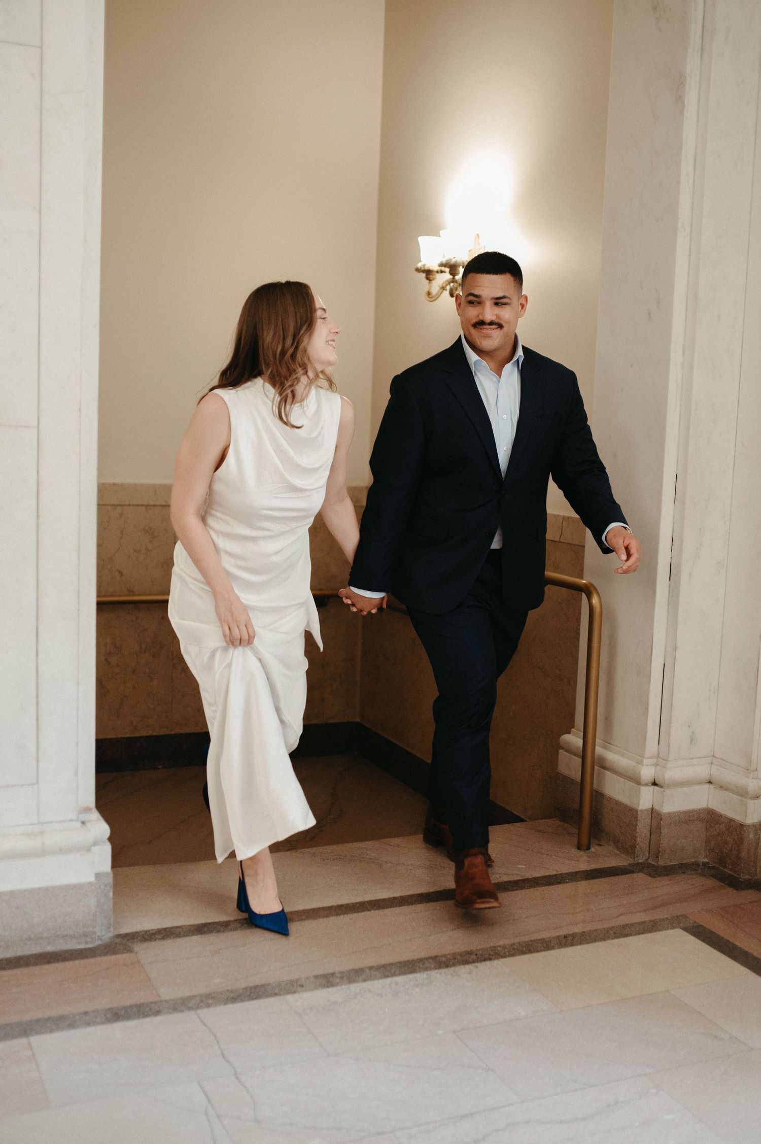 A couple in formal attire walking through an elegant hallway with marble columns.