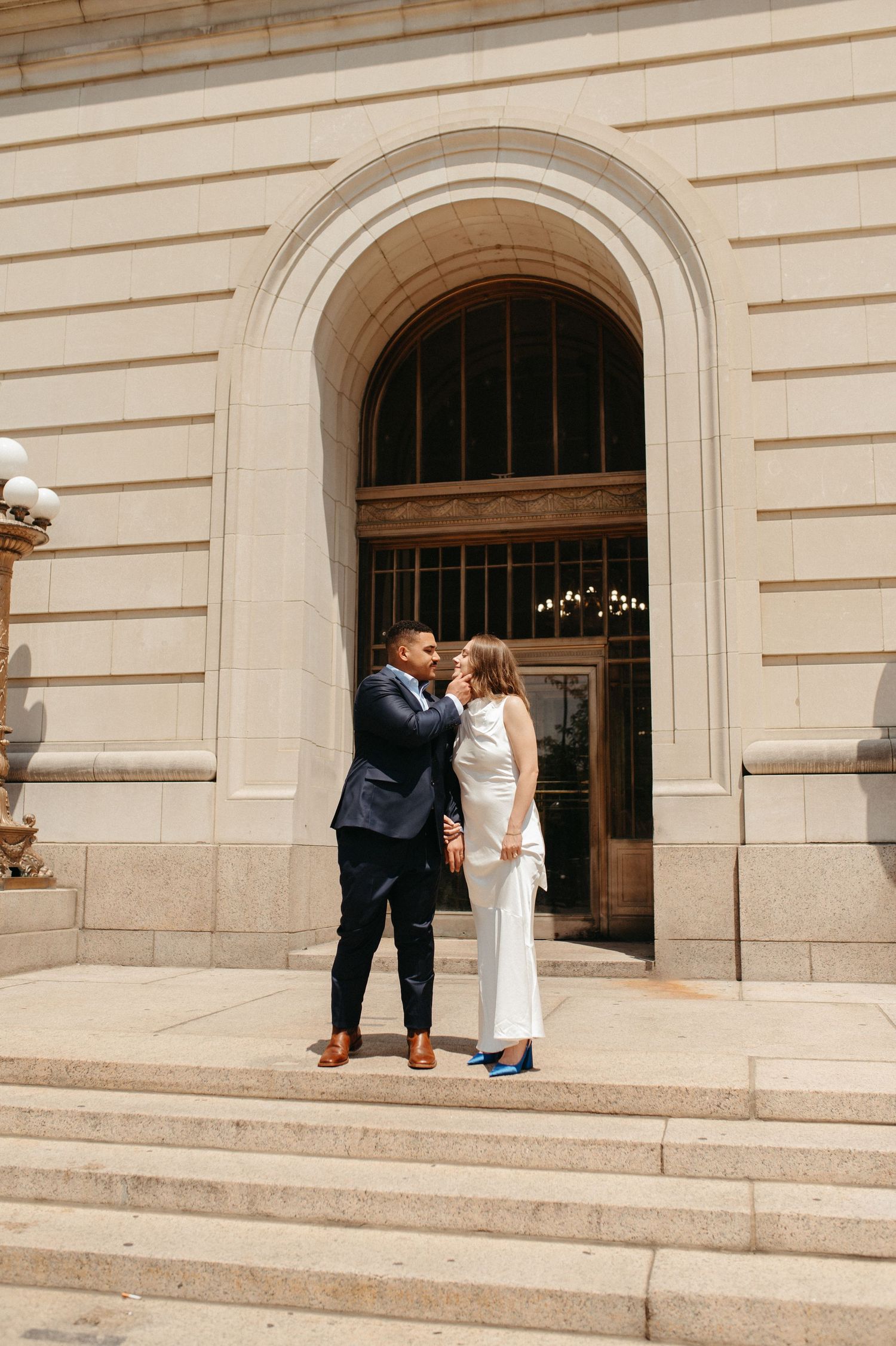 Wedding couple standing on stone steps in front of grand arched entrance to historic building.