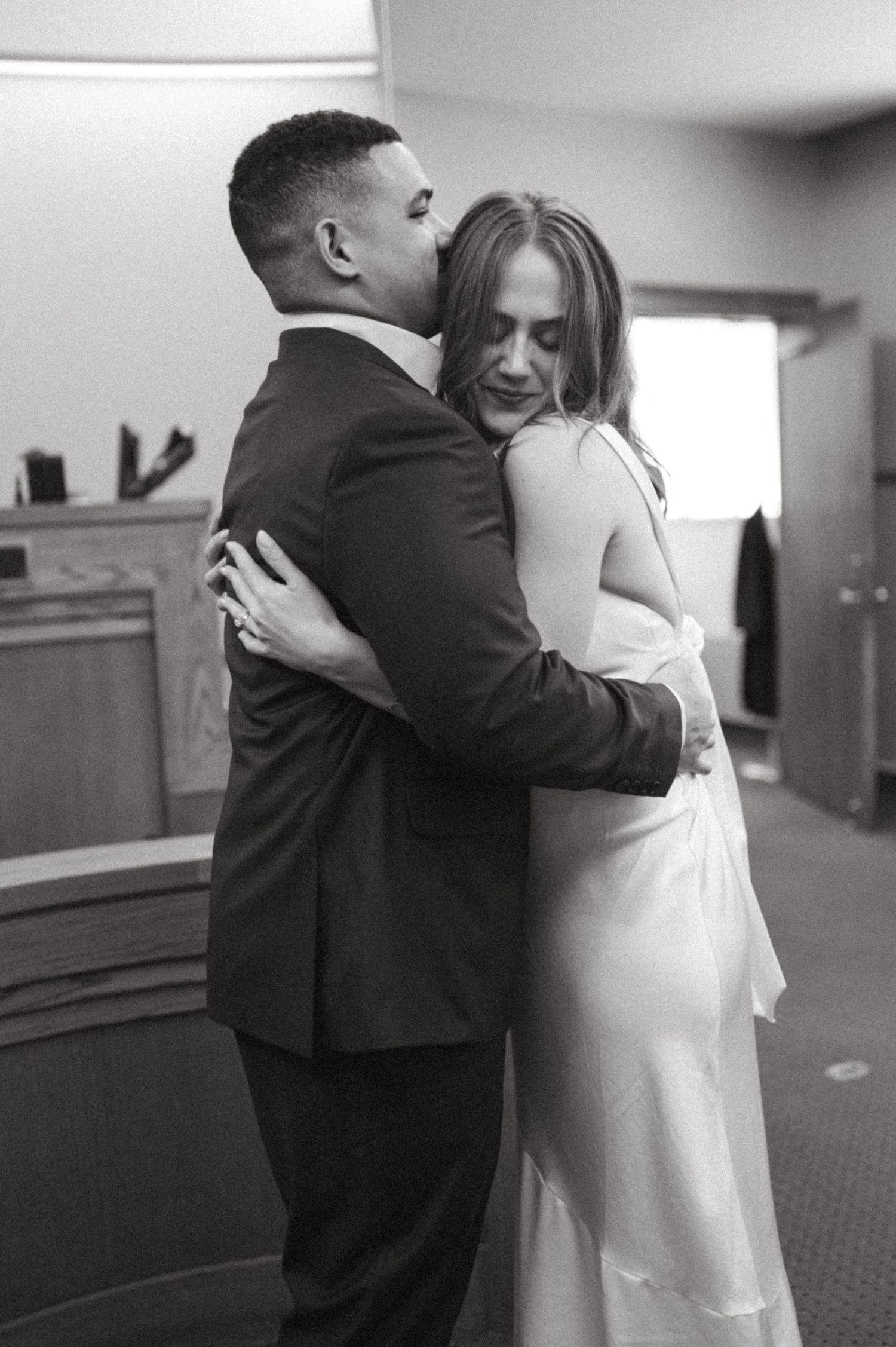 A tender embrace between a couple at an indoor venue, with the groom in a dark suit and bride in a white dress.