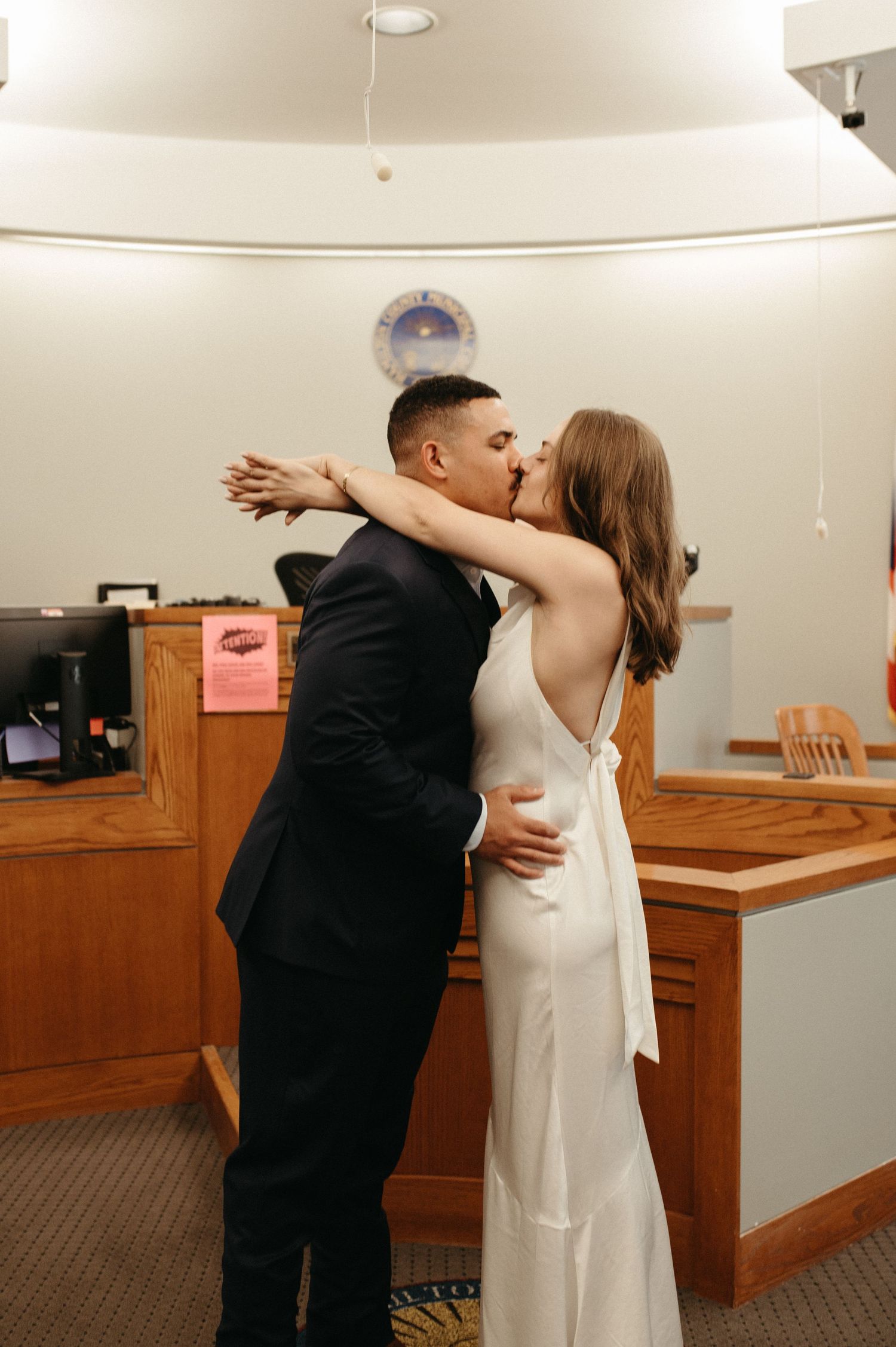 A romantic embrace between newlyweds in formal attire inside a courthouse setting.