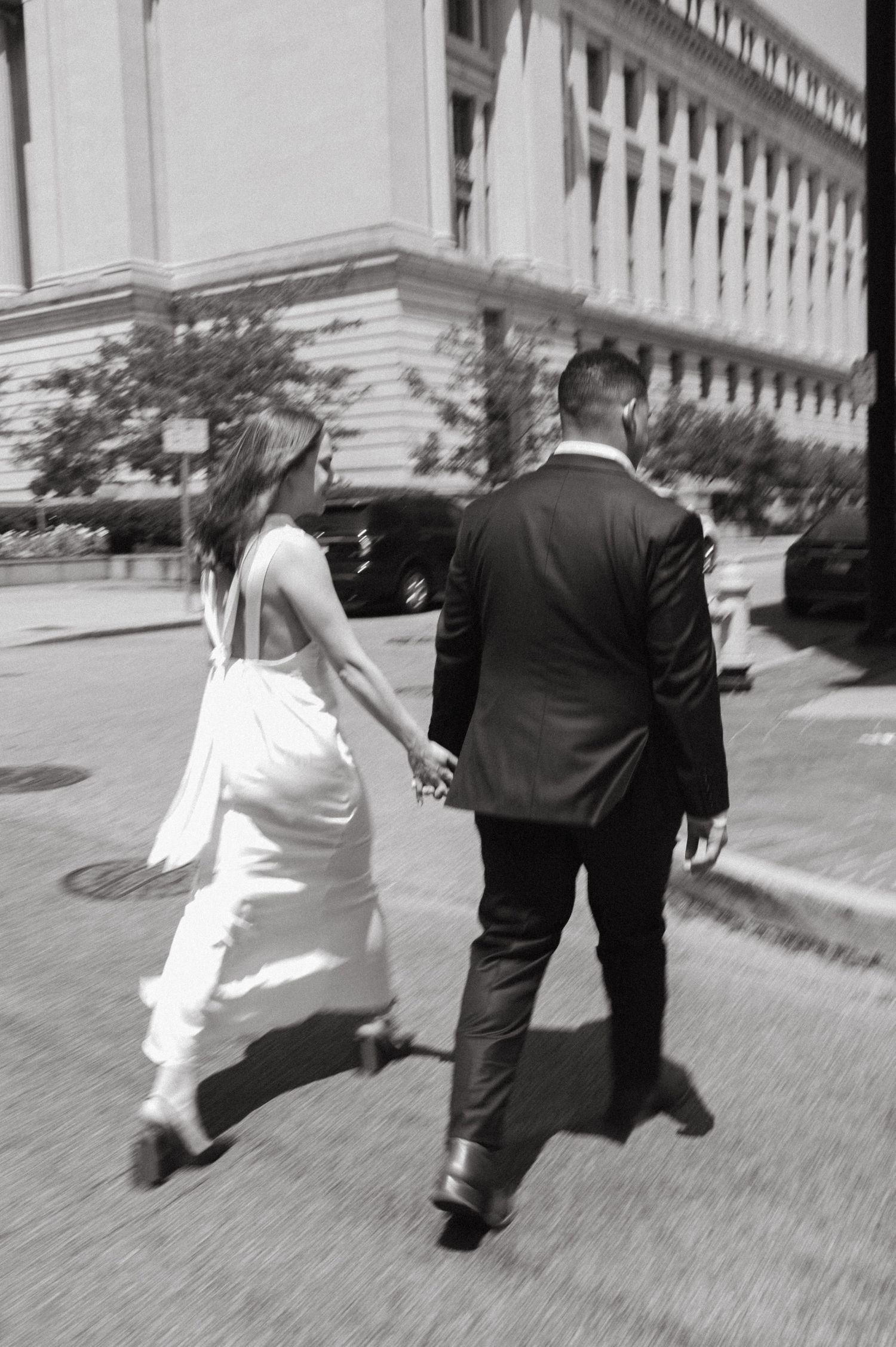 A newlywed couple walks hand in hand down a city street past a grand building.