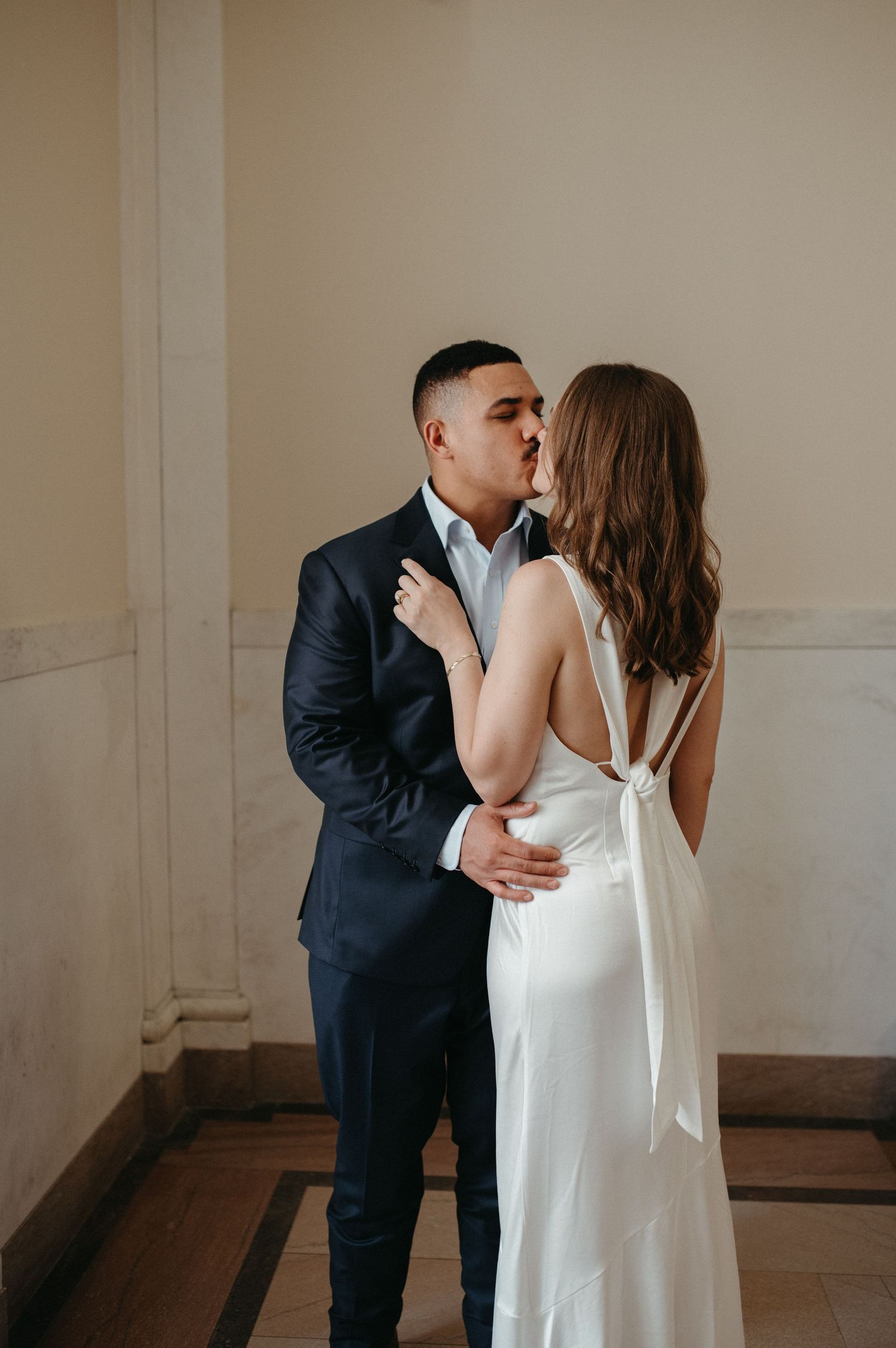 Romantic couple in formal attire share tender moment against beige wall.
