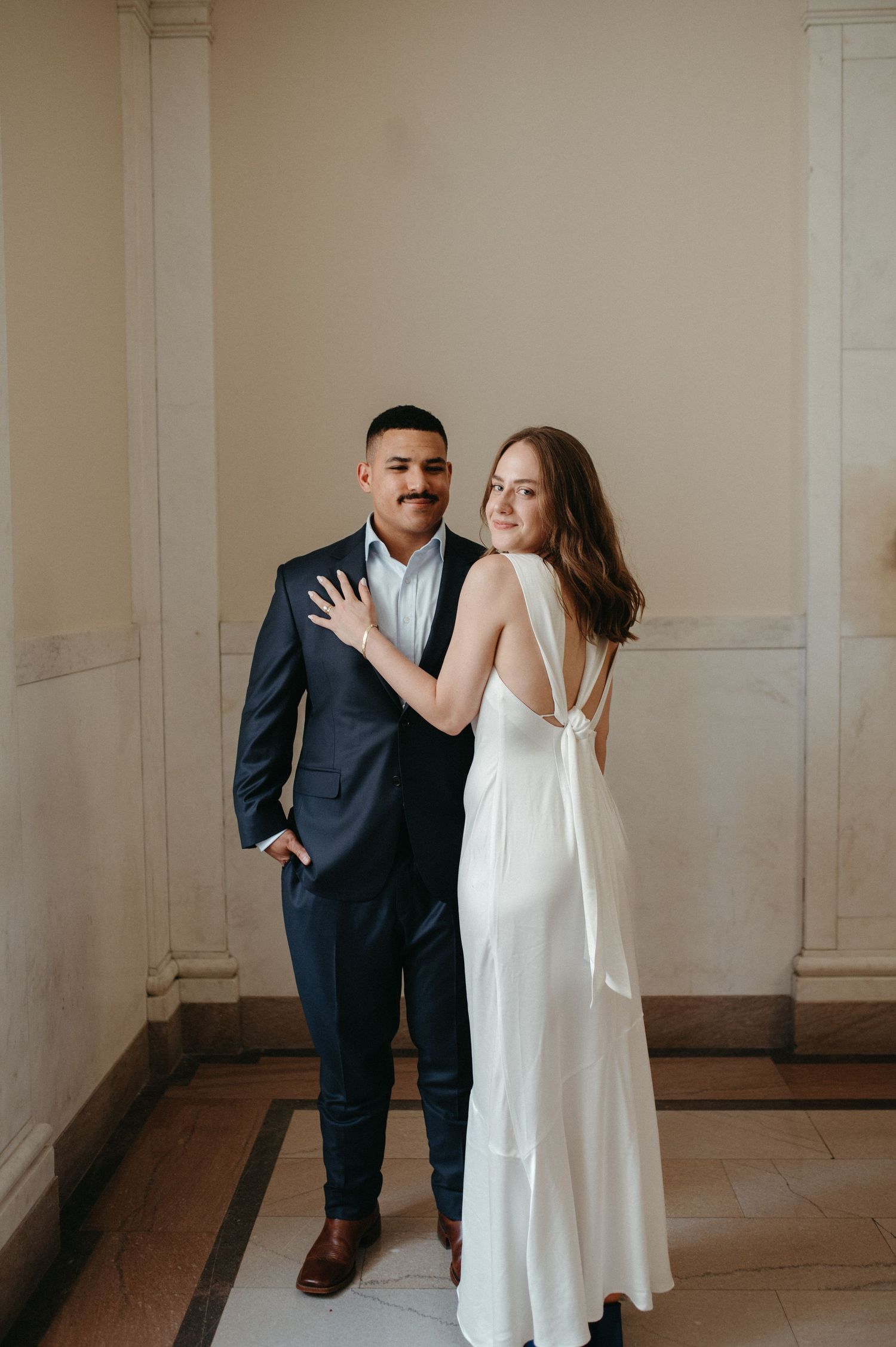 A couple poses together in formal attire against a neutral wall with classic architectural details.