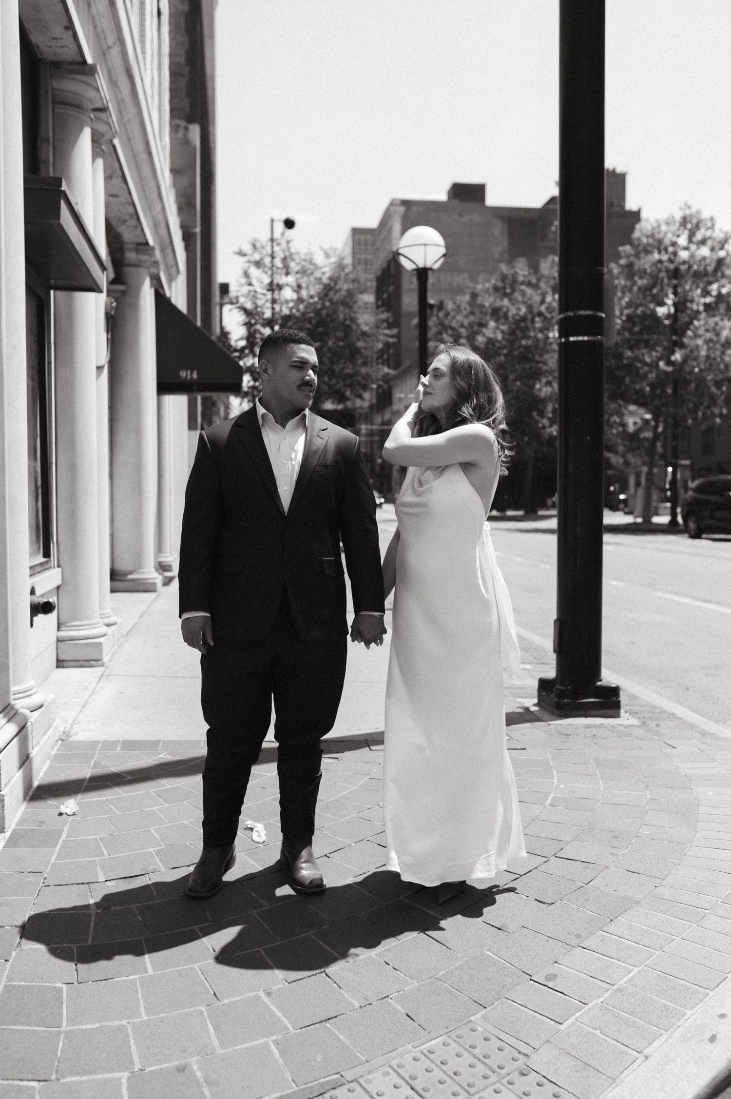 Wedding couple shares a lighthearted moment on a city sidewalk with street lamps and buildings in background.