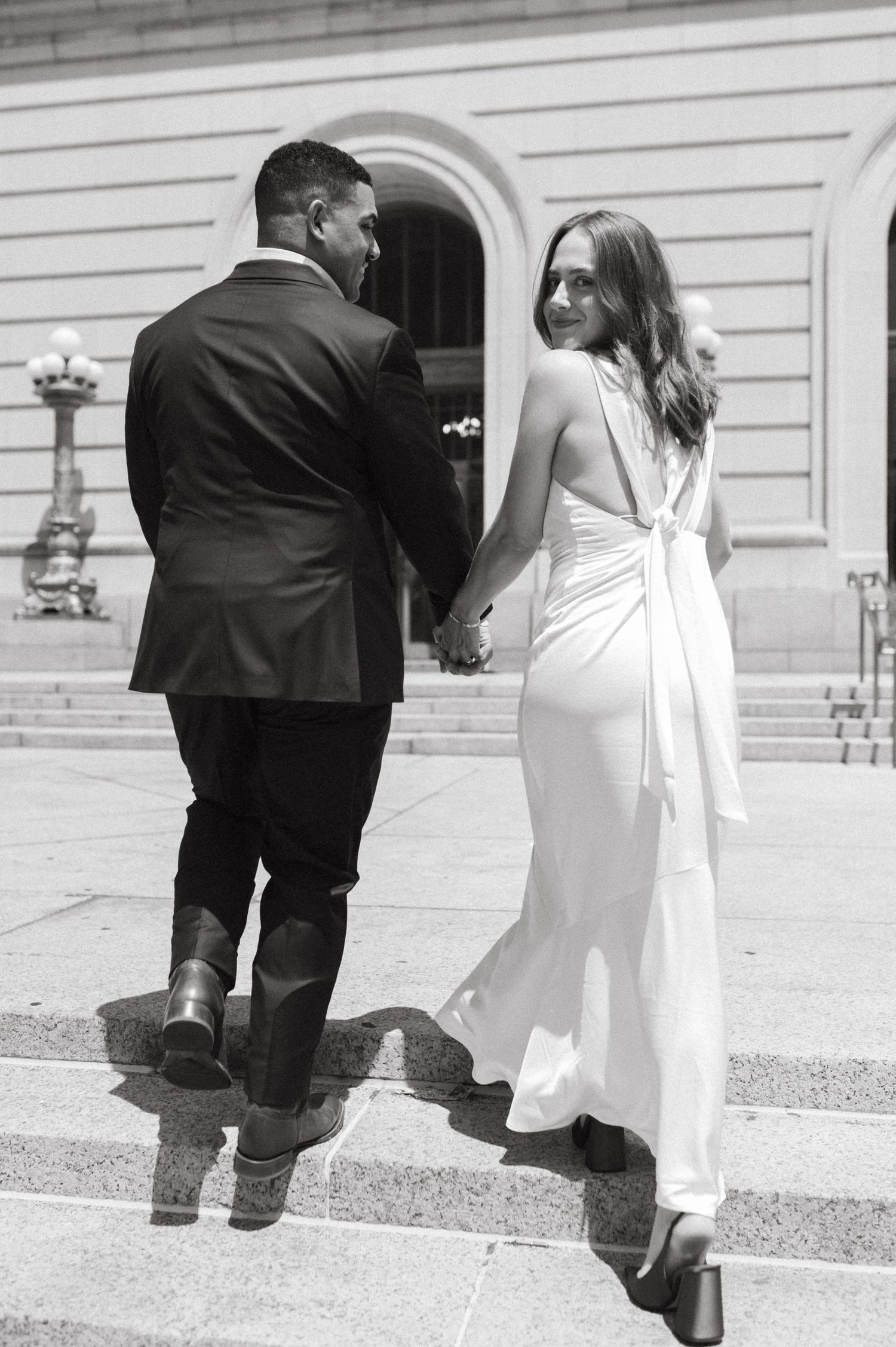 Black and white photo of couple ascending stairs in wedding attire, with bride looking back over shoulder.