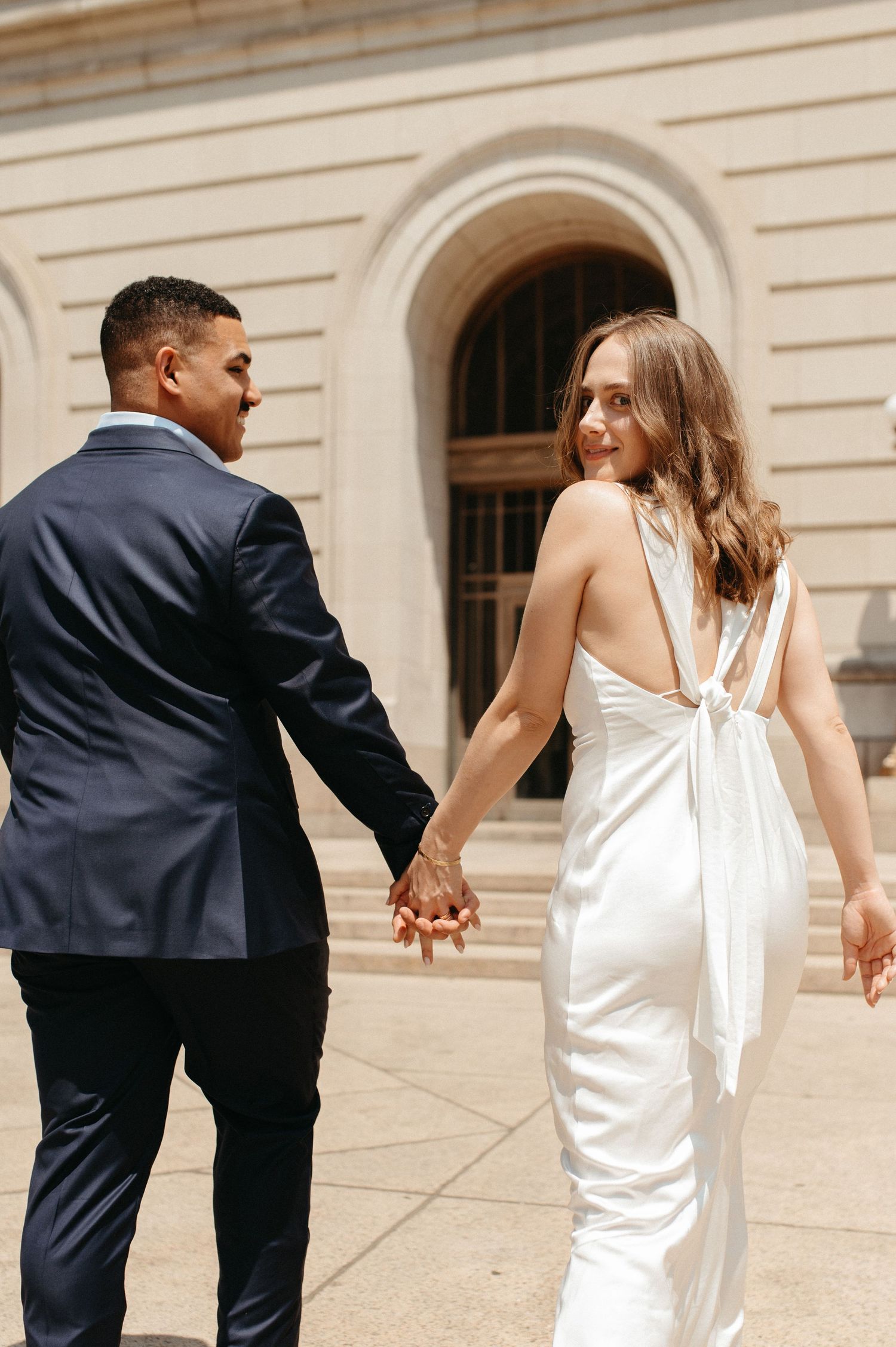 Couple in wedding attire holding hands while walking near classical building with arched doorway.