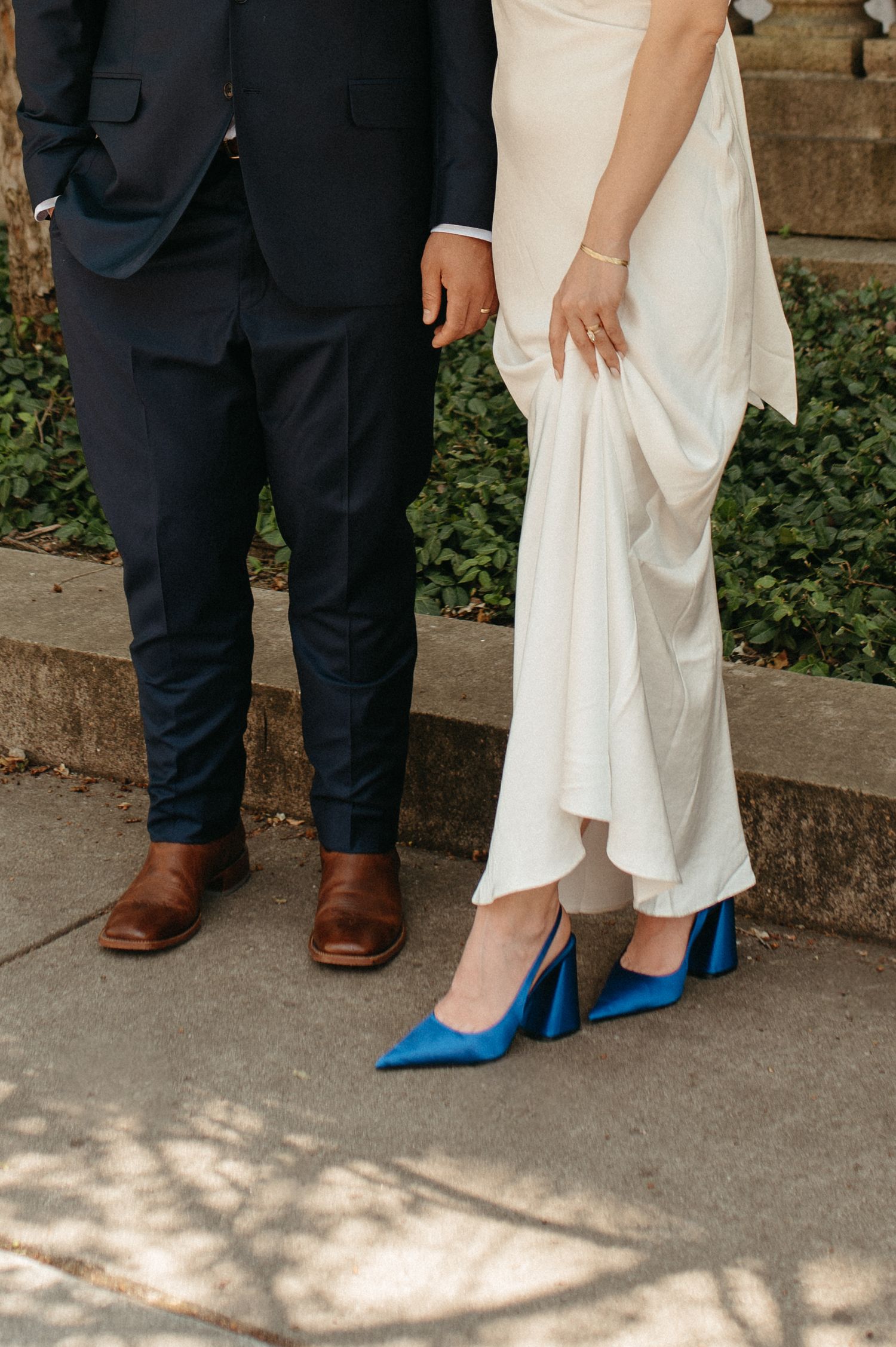 Close-up of bride in white dress wearing vibrant blue heels next to groom in dark suit with brown dress shoes.