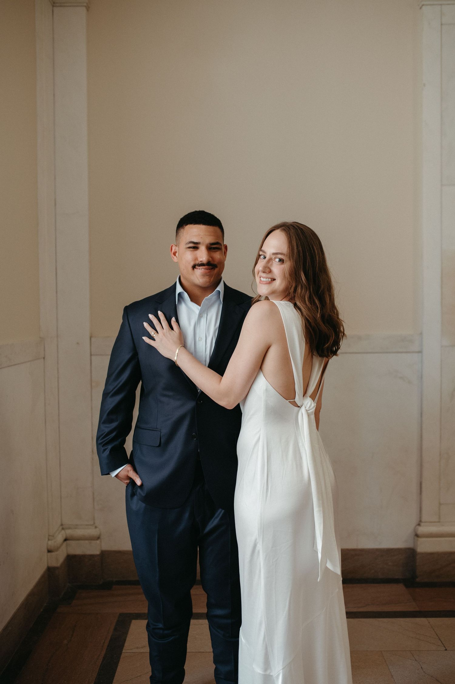 Elegant couple poses in formal attire against neutral wall with bride in white dress showing bow detail.