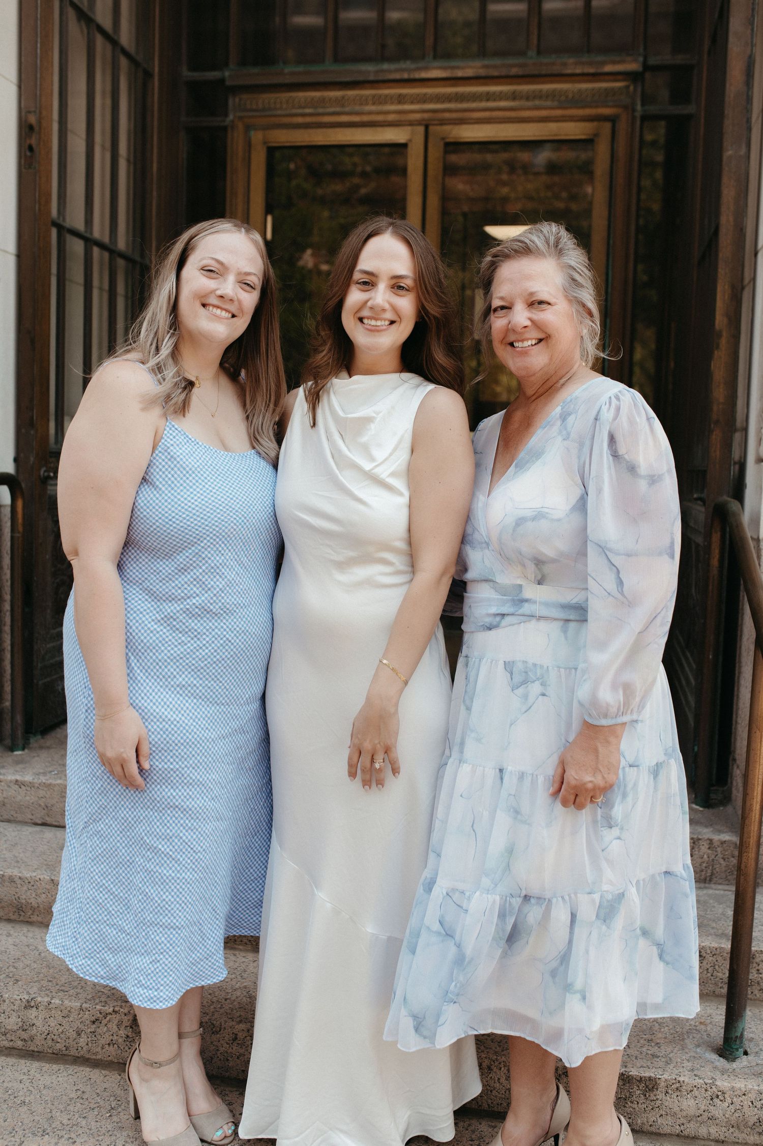 Three women in pastel dresses smile together on building steps.