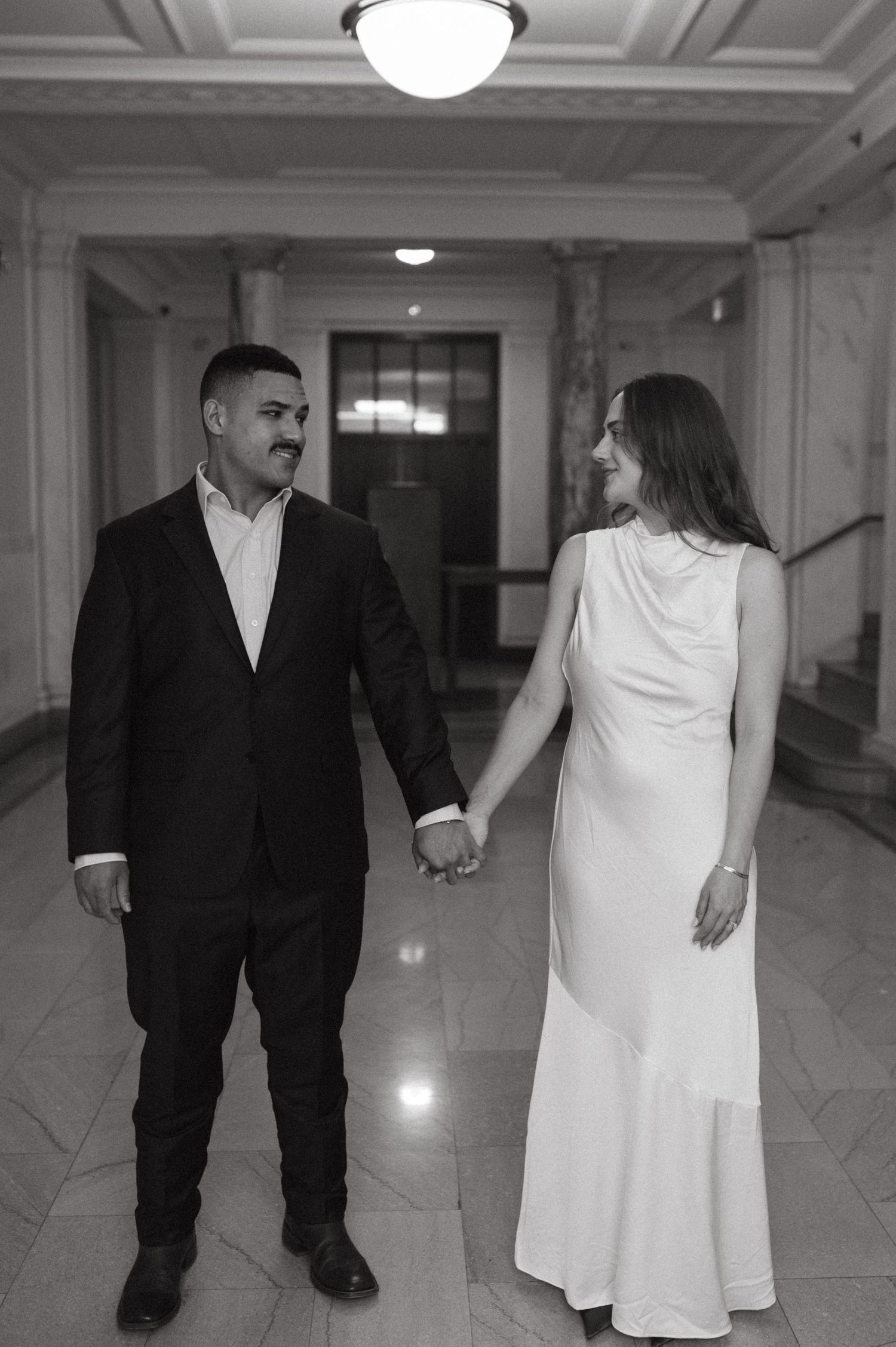 A couple holds hands while walking down an elegant hallway with marble columns and ornate ceiling fixtures.