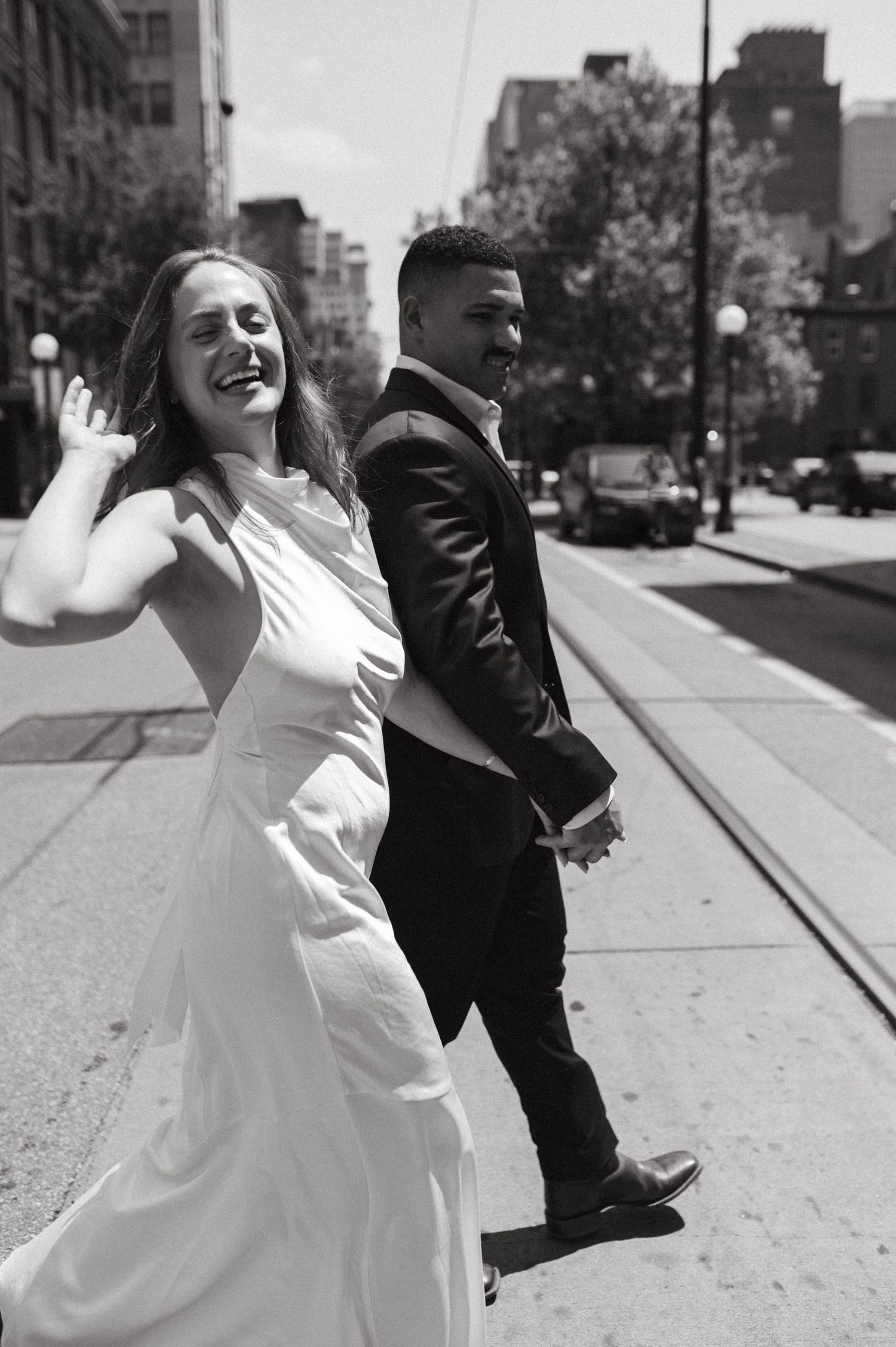 A couple shares a playful moment on a city street, with the bride in white and groom in black walking together.