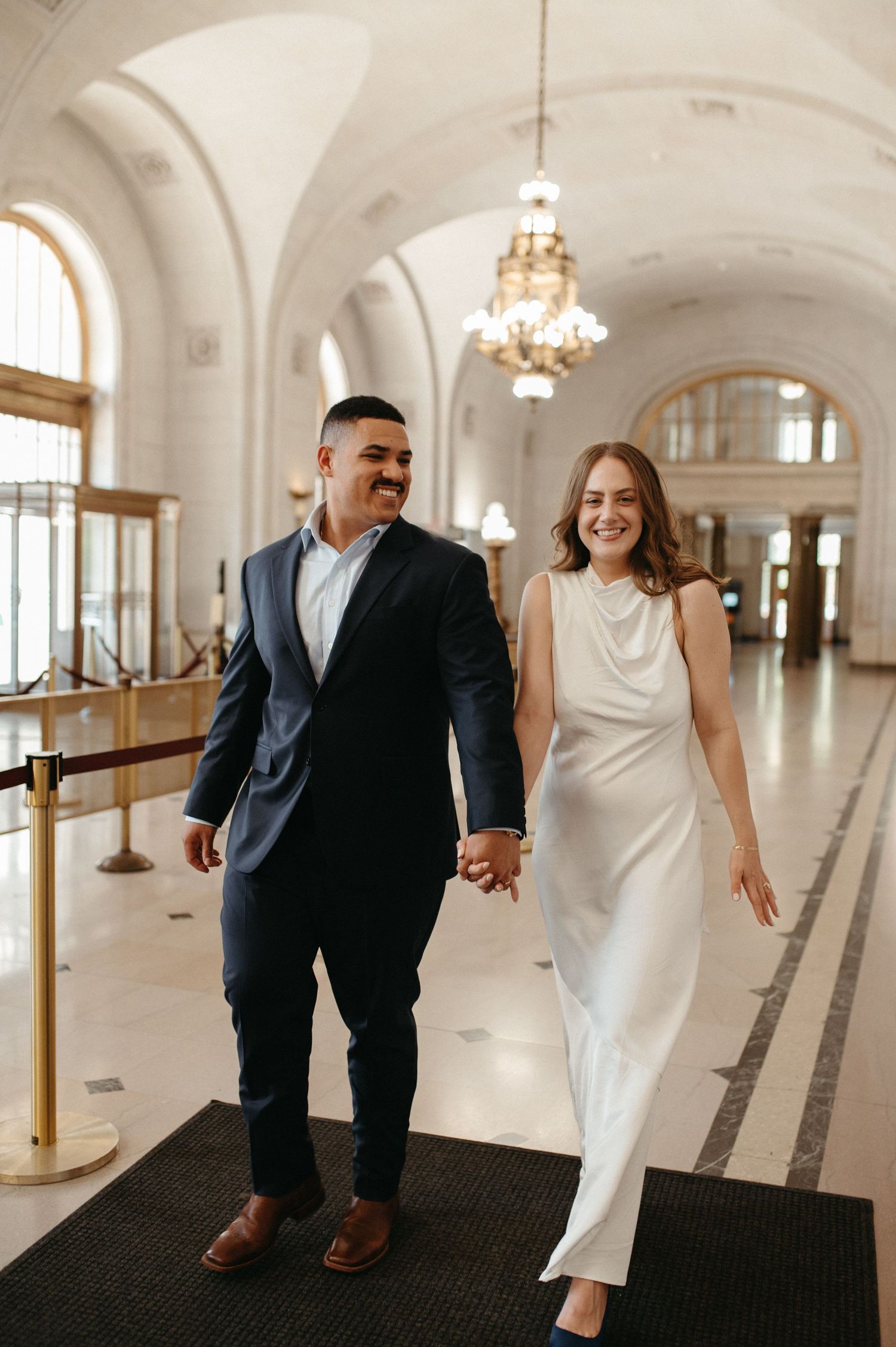 Happy couple walks hand in hand through elegant hallway with vaulted ceilings.