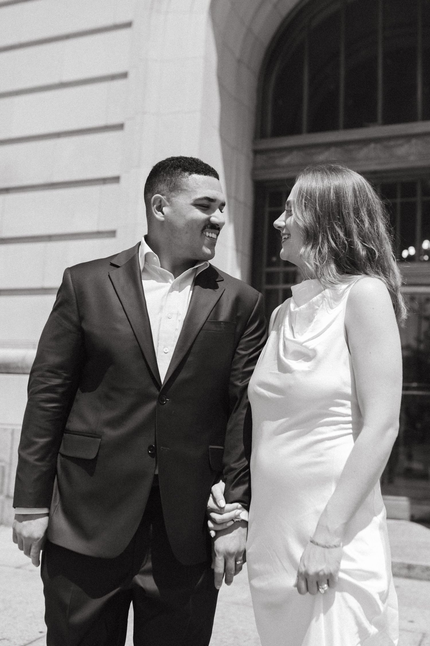 Black and white portrait of couple in wedding attire sharing a joyful moment outside historic building.
