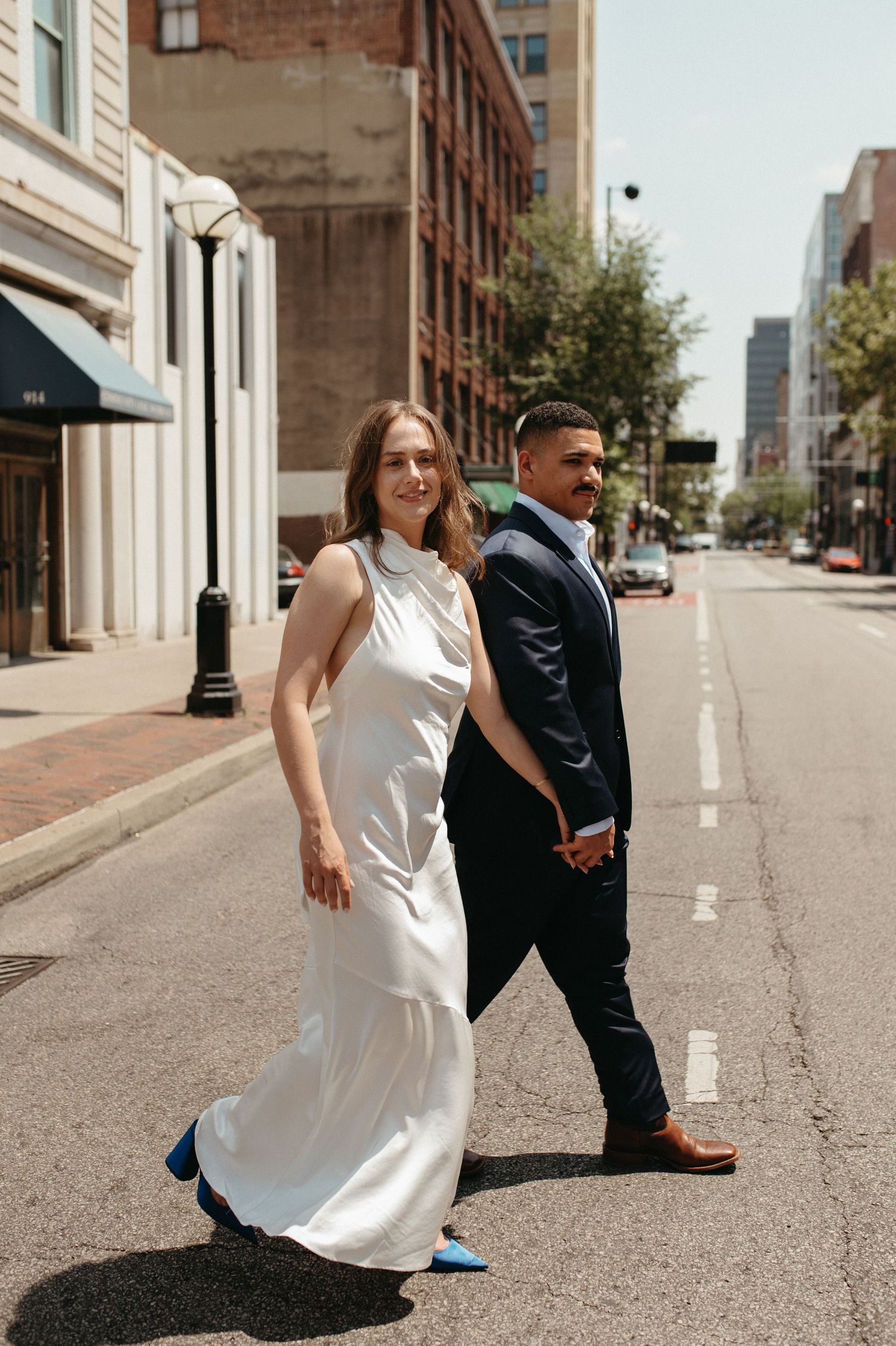 Stylish wedding couple posing on urban street with historic buildings in background.
