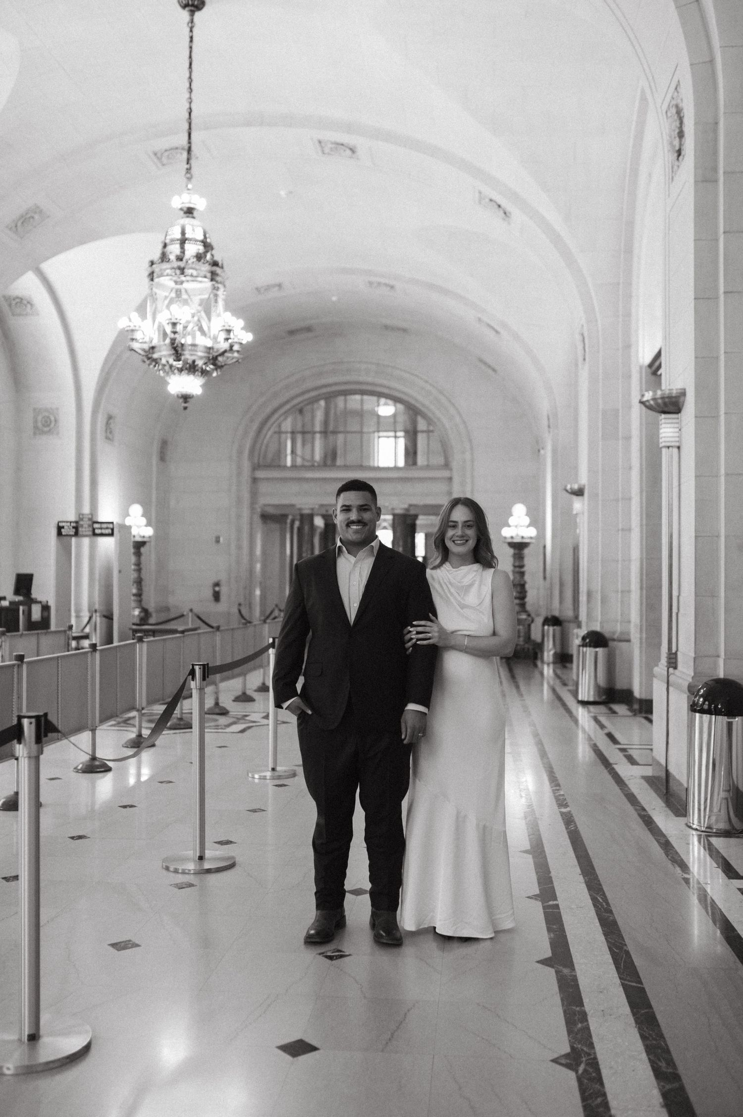 Couple walks through elegant hallway with chandelier and arched ceiling.