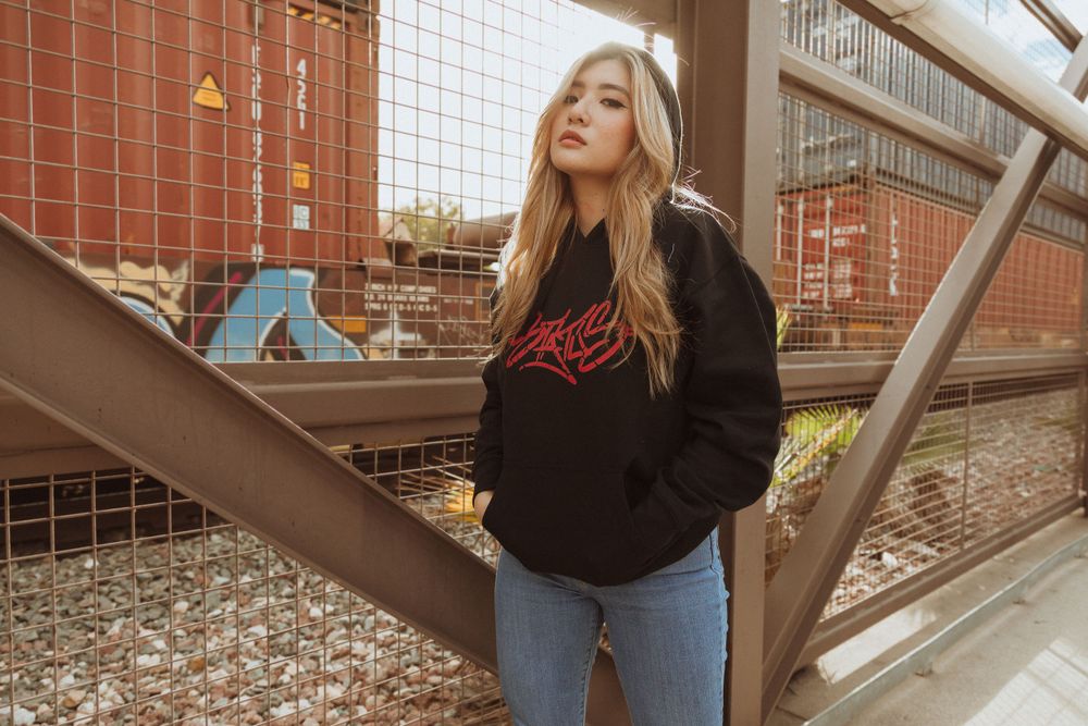 Streetwear fashion model in black hoodie and jeans poses against urban brick backdrop.