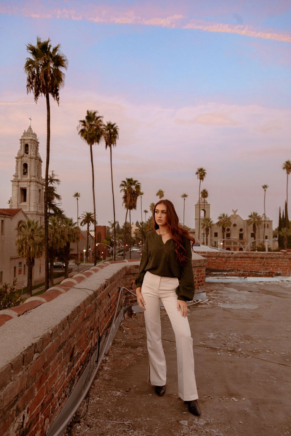 Woman in black sweater and white pants stands on historic building rooftop at dusk.