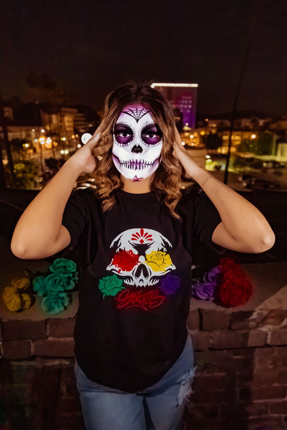 Sugar skull makeup on rooftop with colorful skull t-shirt against night cityscape.