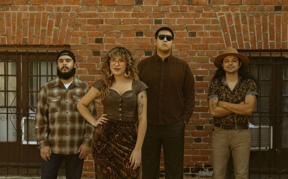 Four musicians in vintage-inspired outfits pose against a brick wall backdrop.