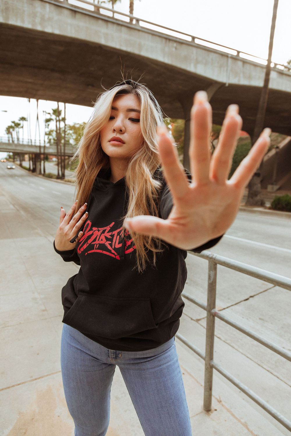 Model in black sweatshirt with red text holds hand up while posing under bridge.