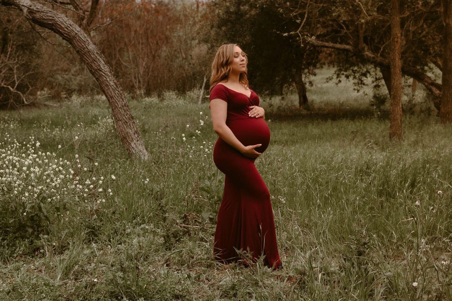 A pregnant woman in a burgundy dress poses in a dreamy outdoor setting with wildflowers.