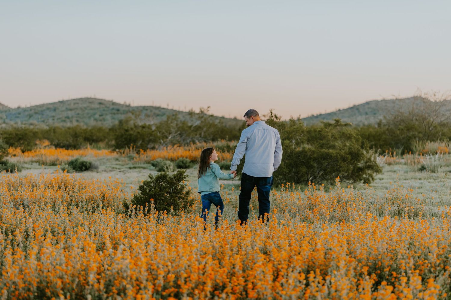 Two people walking together through a field of golden wildflowers at sunset.