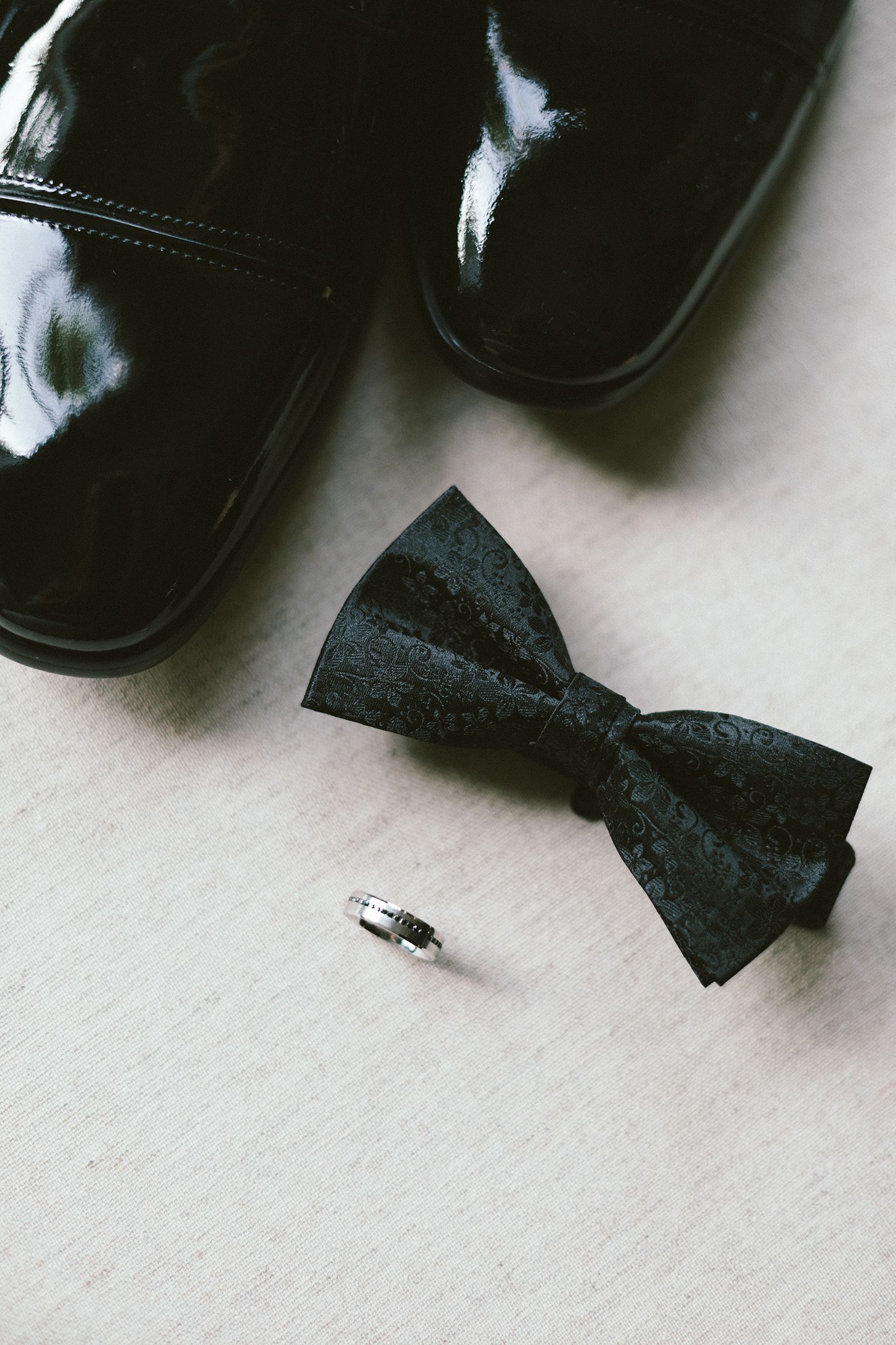 Close-up of black formal shoe tip with bow tie and ring on white surface.