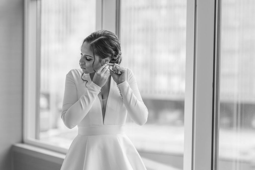 Black and white photo of elegant bride putting on earrings by window.