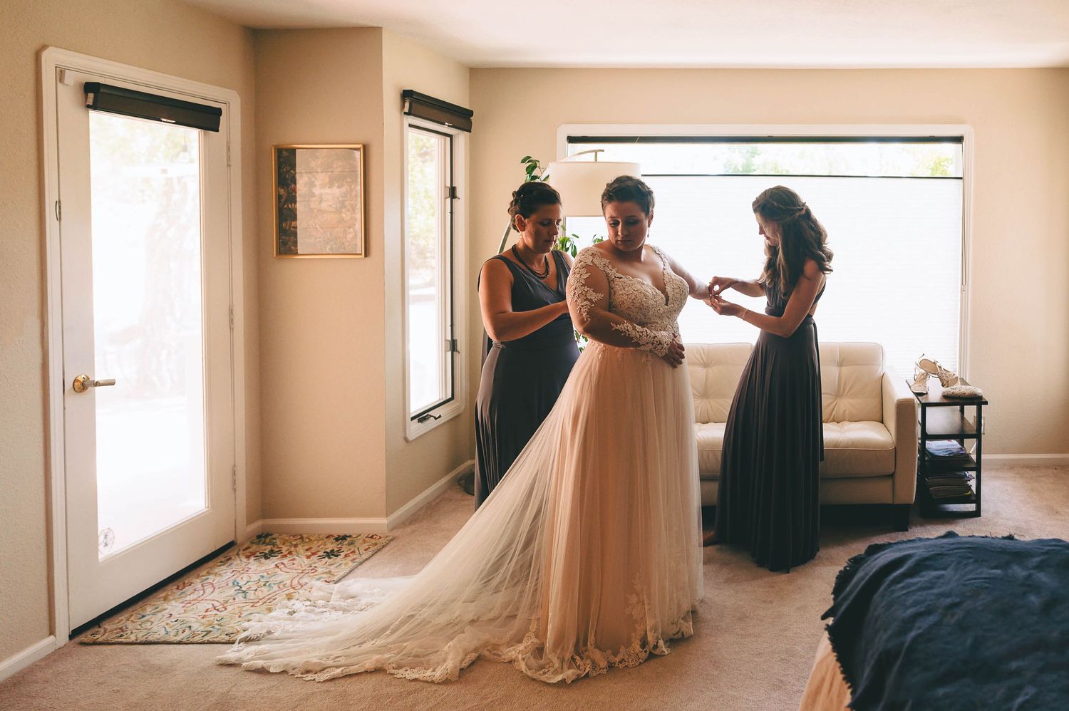 Wedding preparations scene with a bride being helped into her dress by two attendants.