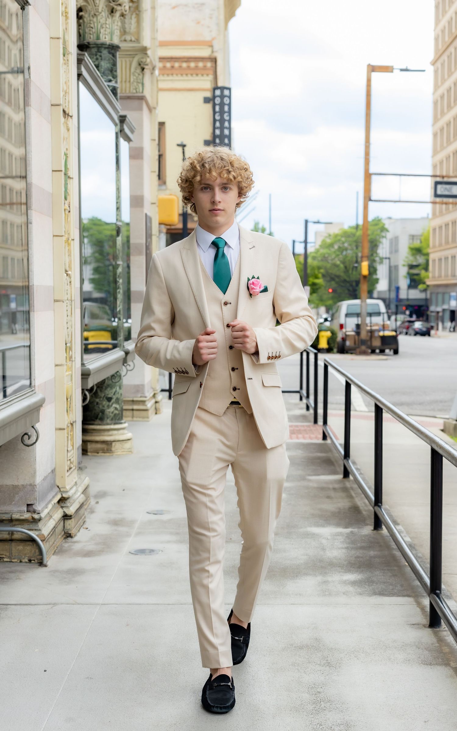 Young person in cream three-piece suit and teal tie walks along urban storefront.