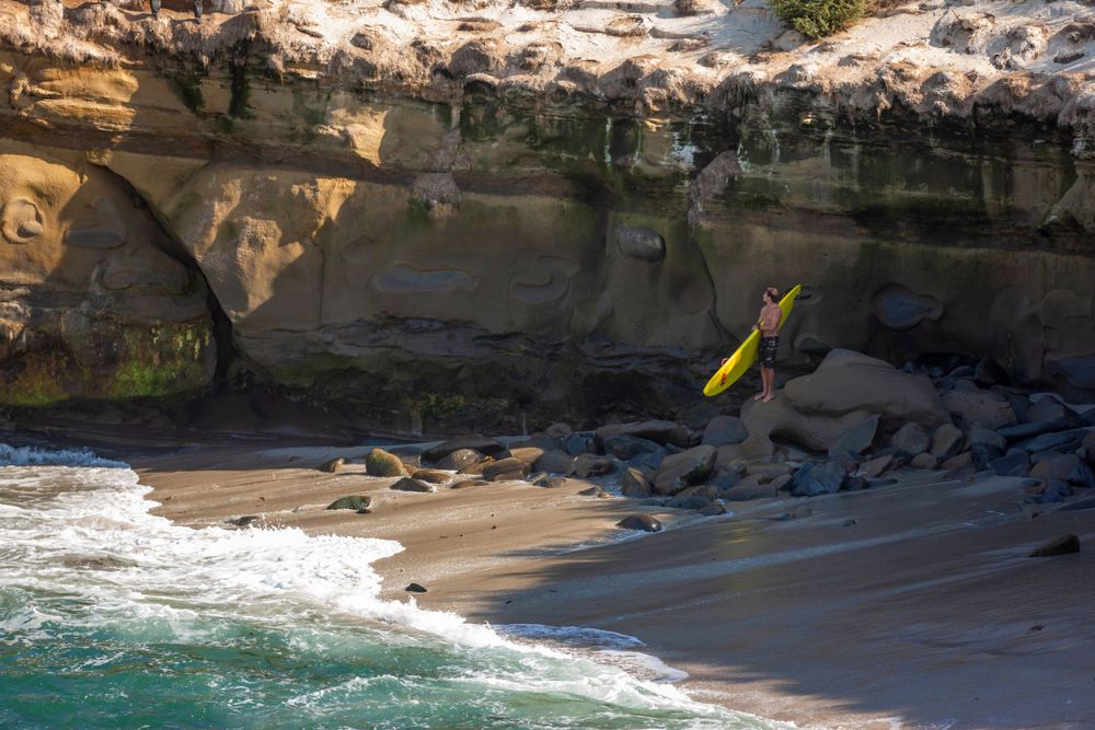 surfer with yellow surfboard standing on rocks under the sandstone cliffs in La Jolla Cove in San Diego, California