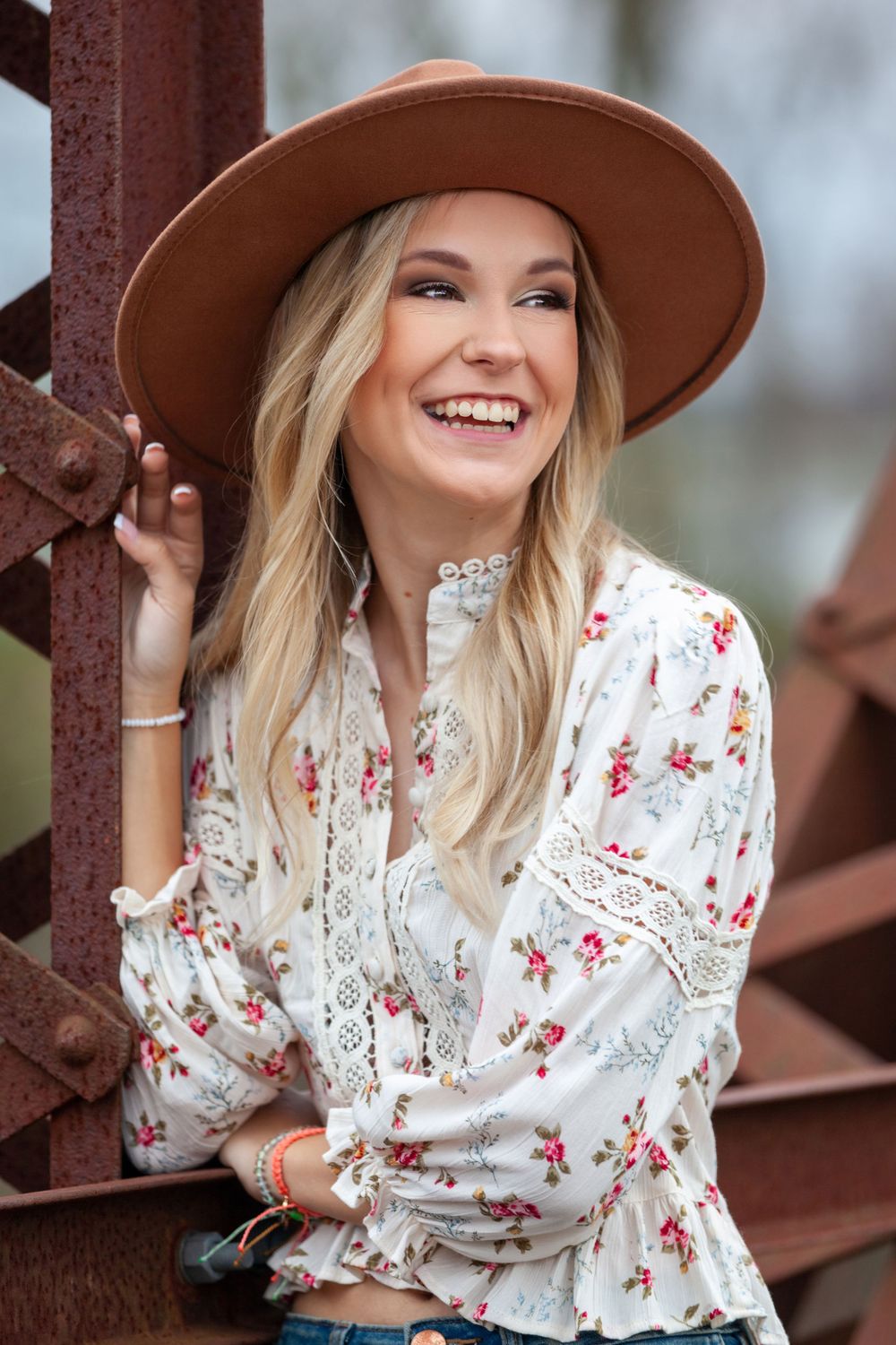 senior girl in brown hat leaned on old metal bridge with big smile looking off to the side on a cloudy day in Judsonia, Arkansas