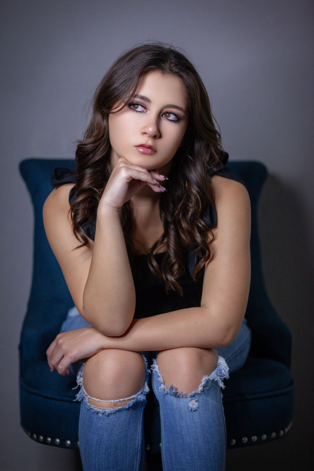 senior girl in ripped jeans sitting with elbows on knees and chin in hand seated in front of plain gray wall in studio in Savannah, Texas