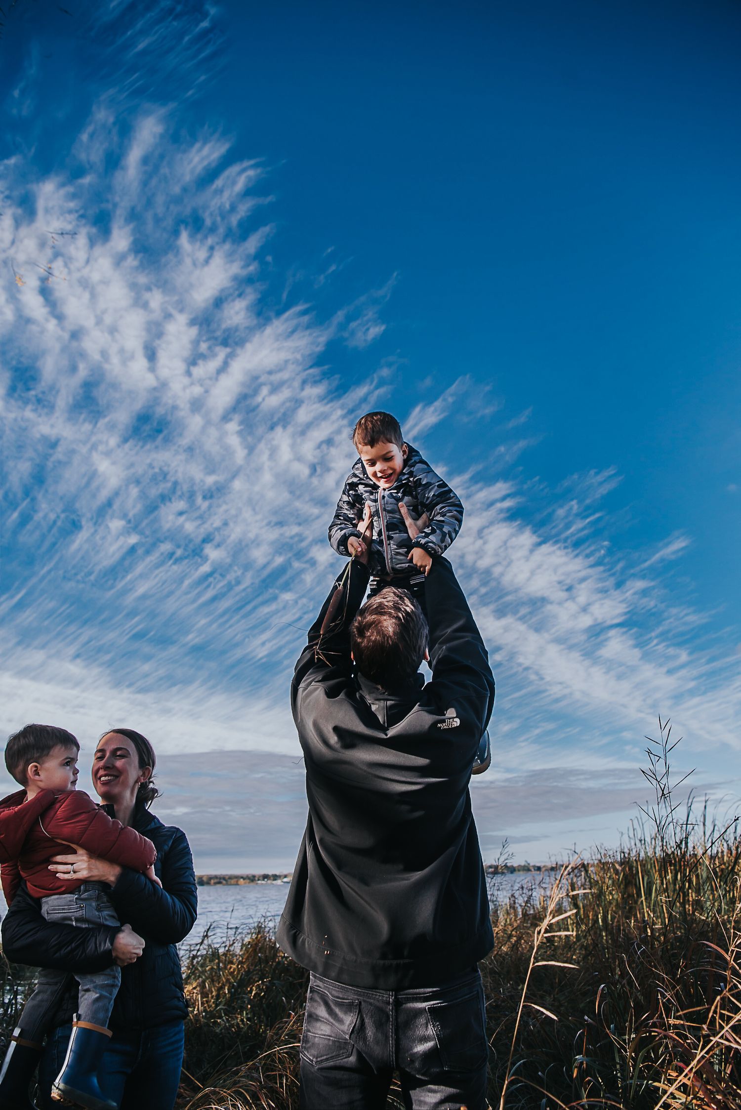 A man holds his child high in the air with blue skies and white clouds behind the.