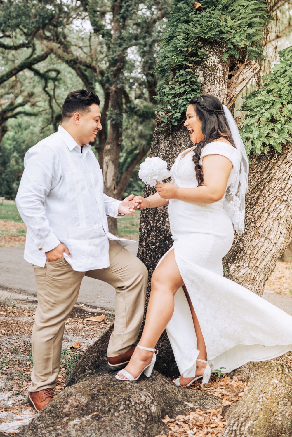 Playful wedding couple dancing and laughing by large tree trunk.