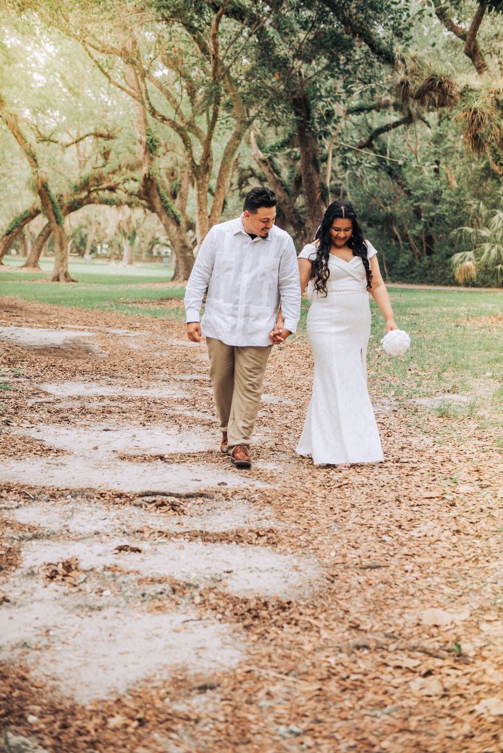 Newlyweds walking down tree-lined path in wedding attire.