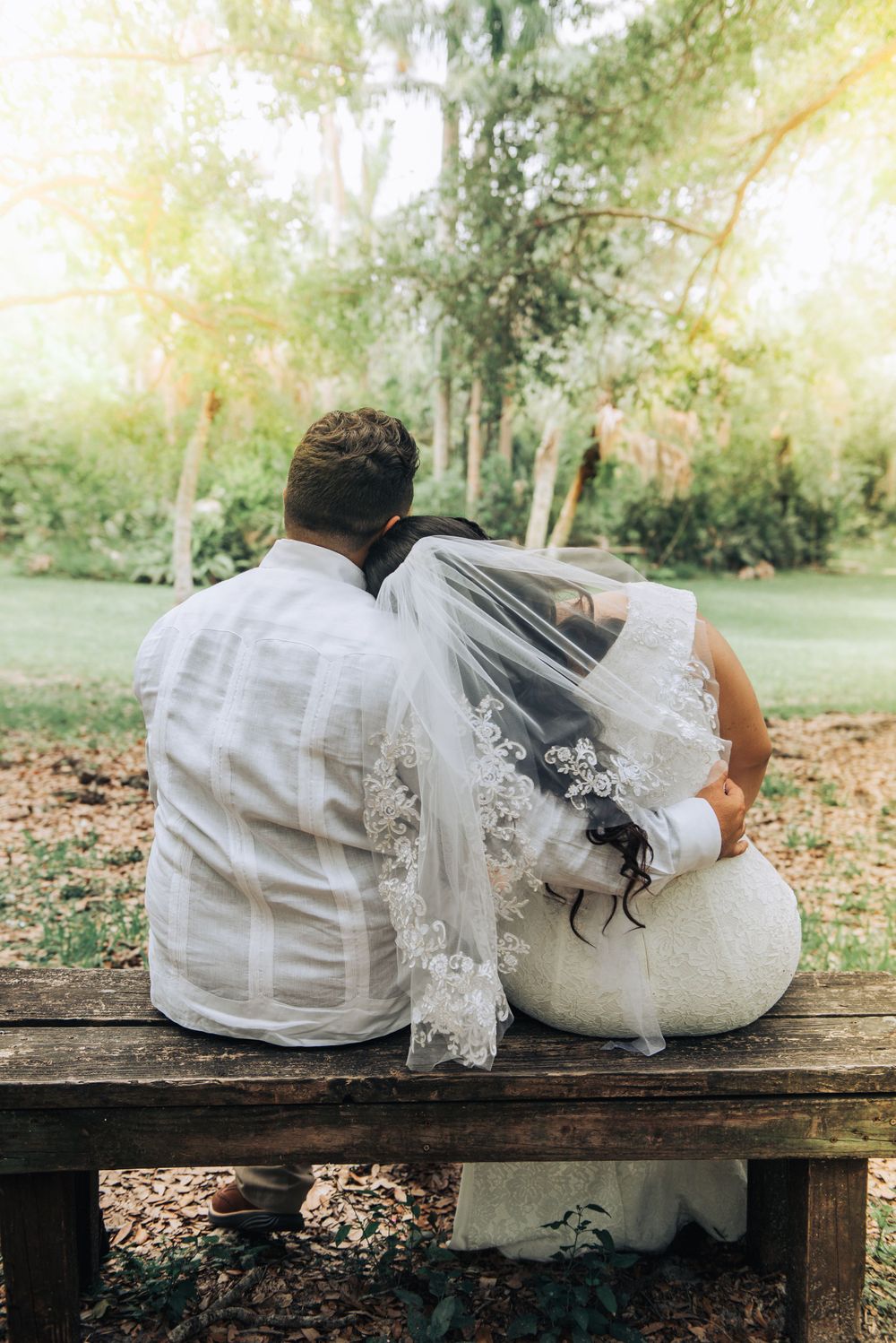 A couple sits on a wooden bench sharing an intimate moment under trees.