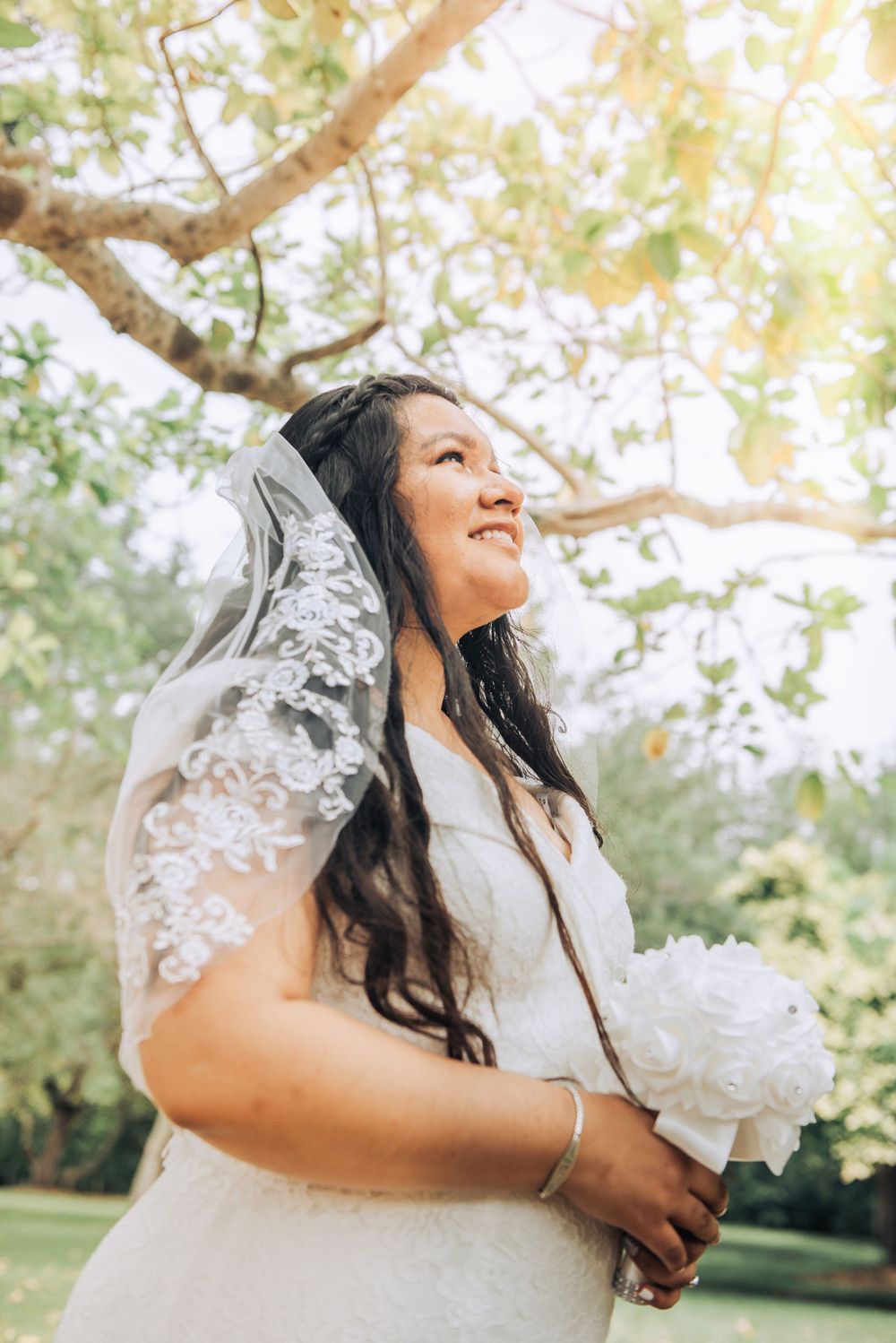 A bride in a white dress and lace veil smiles while holding a bouquet in a sunlit garden.