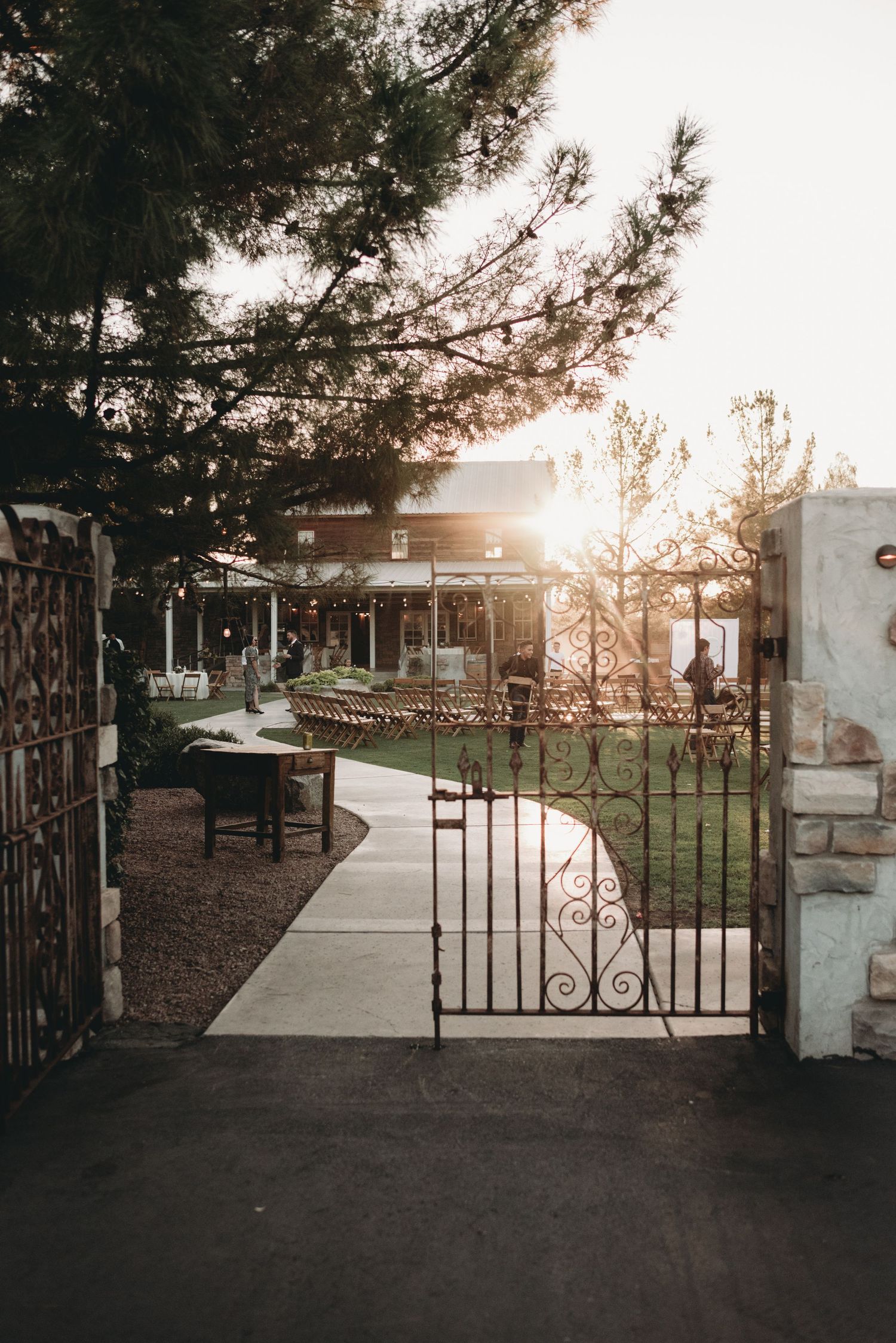 Wrought iron gates frame a garden path at sunset with trees in the background.