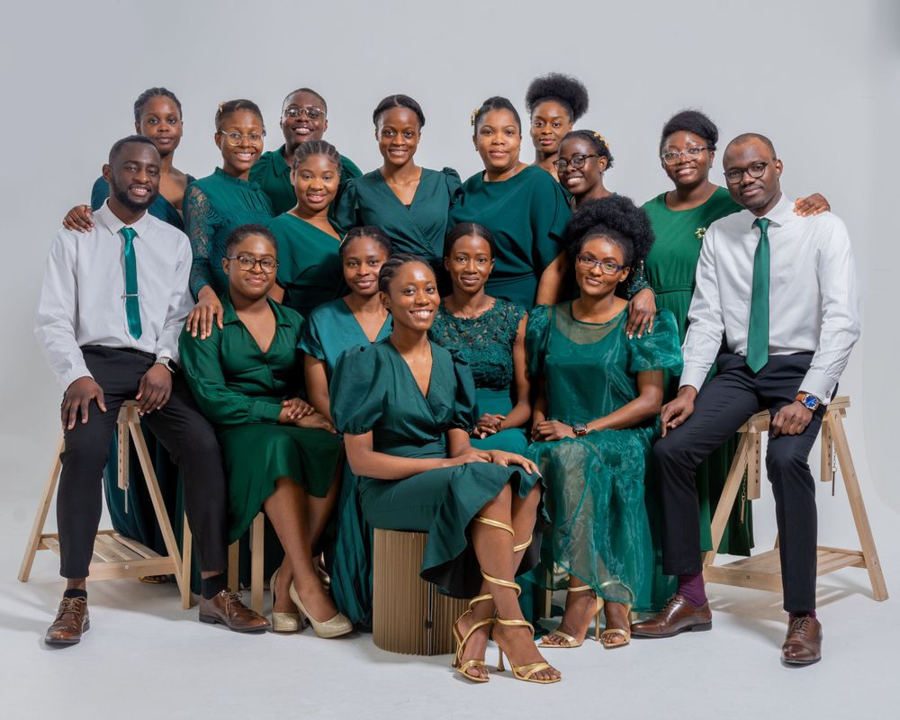 Large group of smiling people in green dresses and white shirts posing for a studio portrait.
