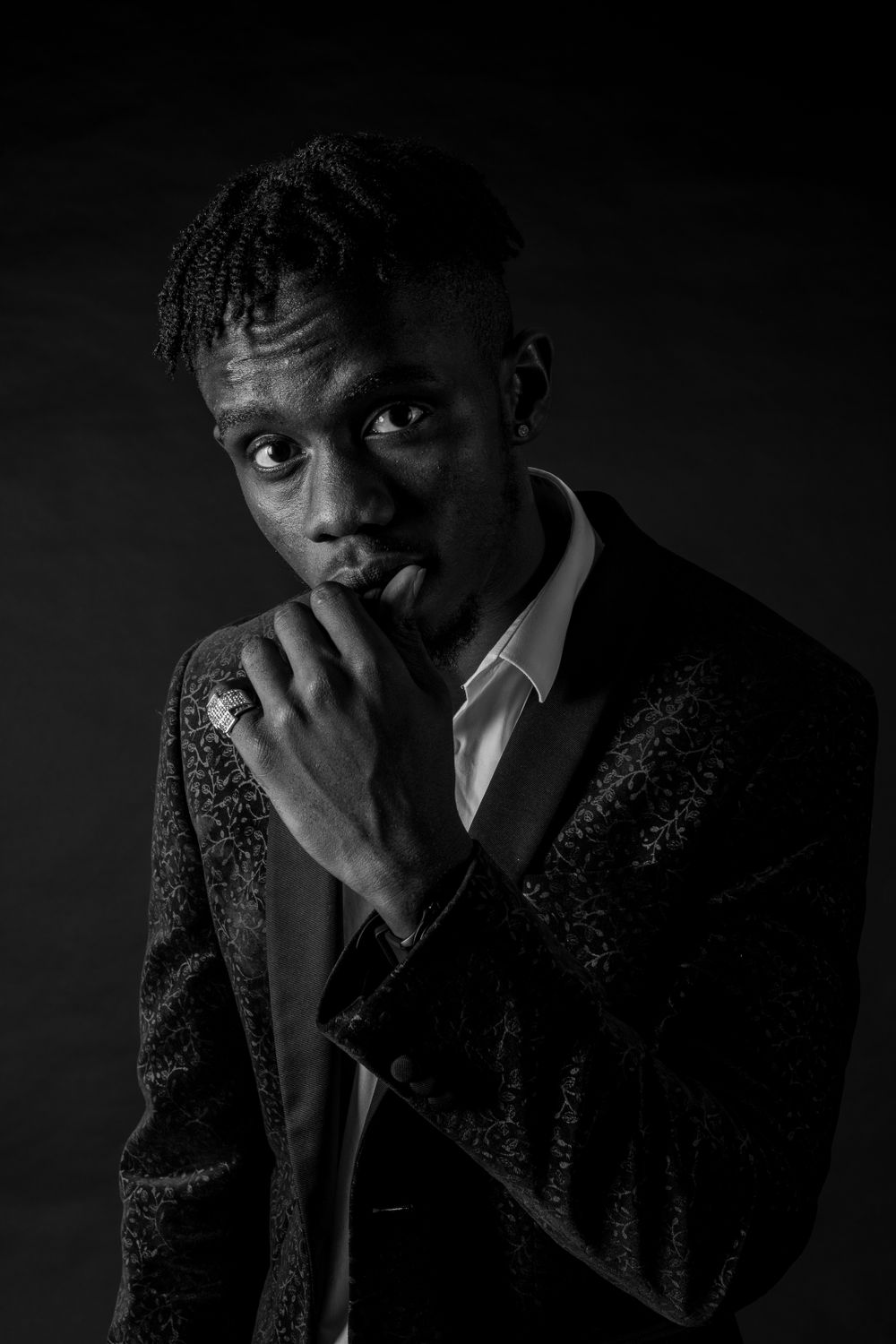 Black and white portrait of young Black man in sequin blazer posing thoughtfully against dark background.
