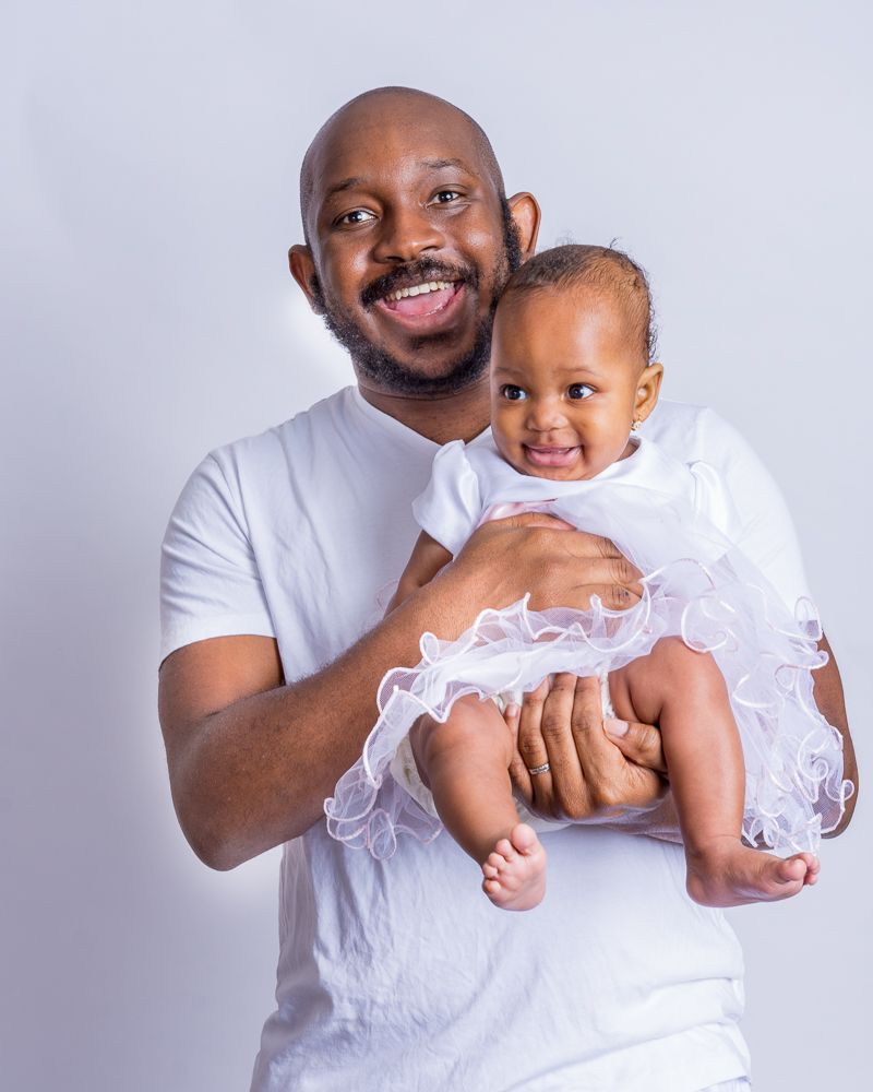 Smiling bald father in white shirt holding happy baby girl in white dress.