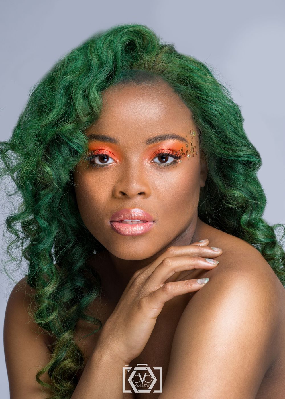 Black woman with bold green curly hair and orange eye makeup posing for a beauty portrait.