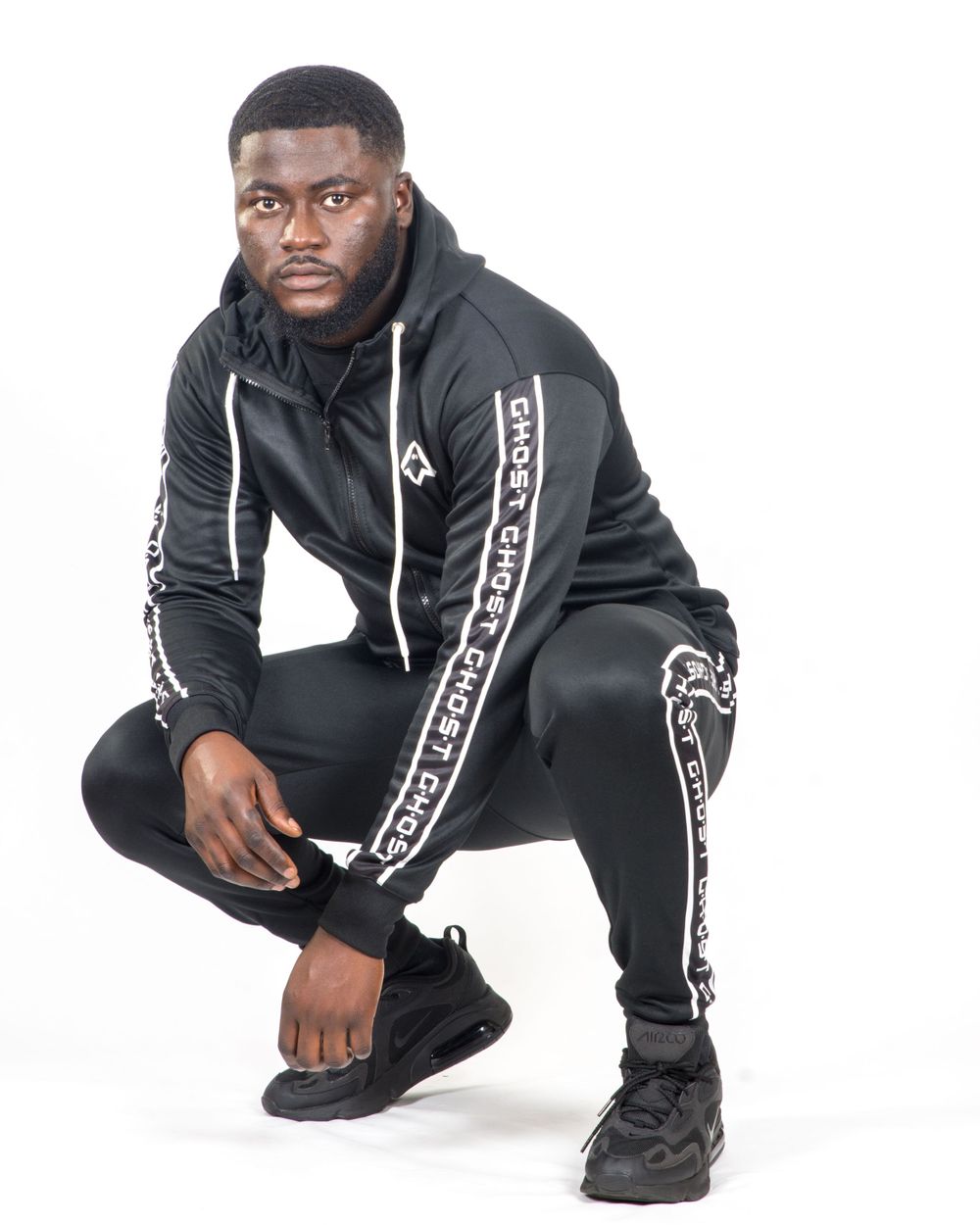 A young Black man crouching in a black Ghost branded tracksuit and black sneakers against a white background.